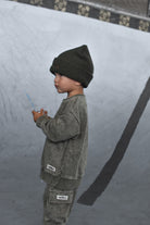 A child standing in profile wearing a dark olive green beanie, a matching washed green sweatshirt, and green cargo pants in a concrete bowl.