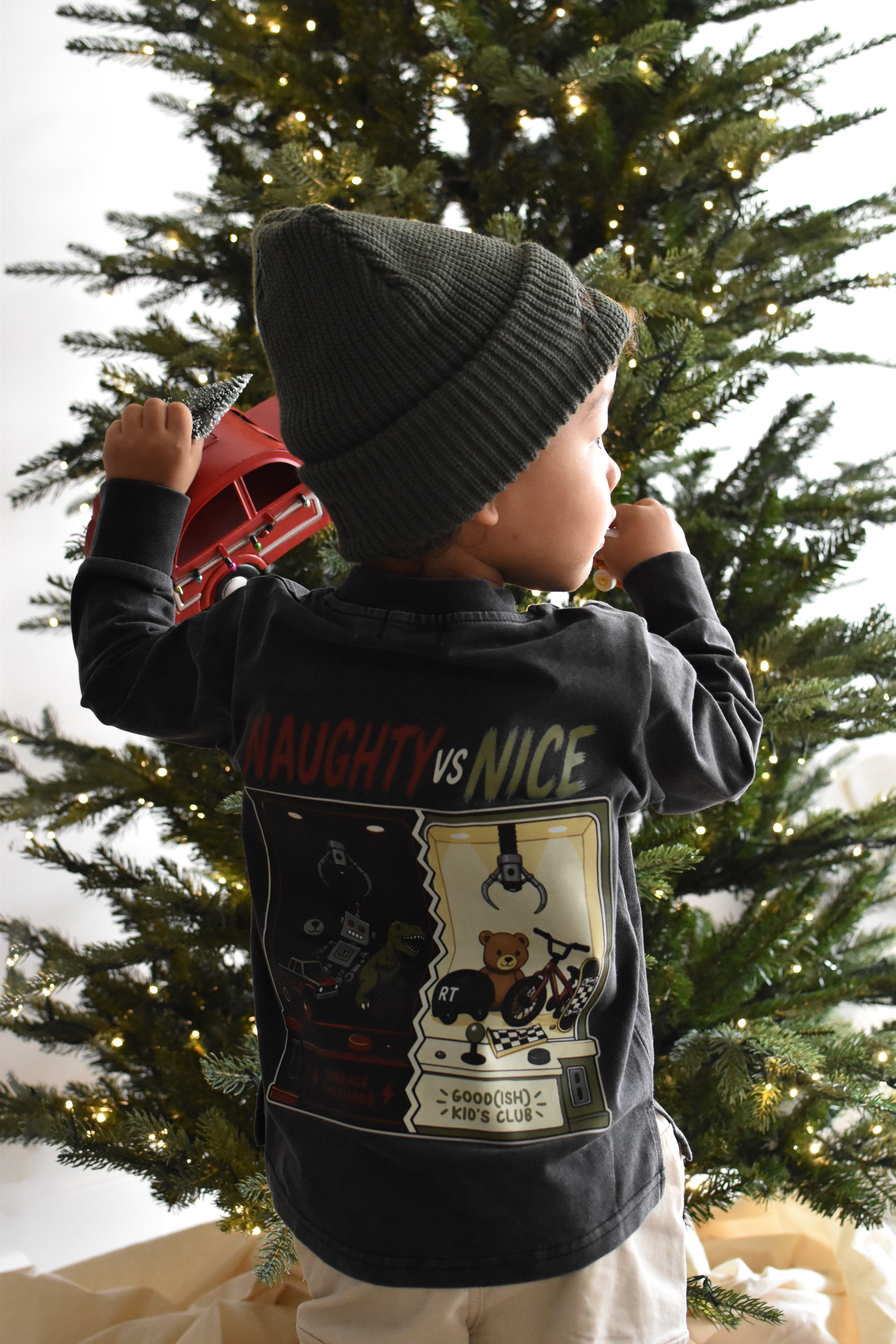 A toddler viewed from behind wearing a dark charcoal gray long-sleeve graphic shirt and a dark knit beanie, standing in front of a decorated Christmas tree.