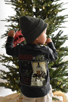 A toddler viewed from behind wearing a dark charcoal gray long-sleeve graphic shirt and a dark knit beanie, standing in front of a decorated Christmas tree.