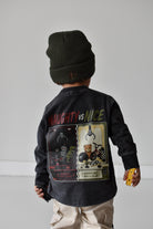 A child shown from the back wearing a dark charcoal long-sleeve graphic jacket with 'NAUGHTY vs NICE' text and a large retro game-style print, paired with a dark green beanie and beige pants against a light gray studio background.
