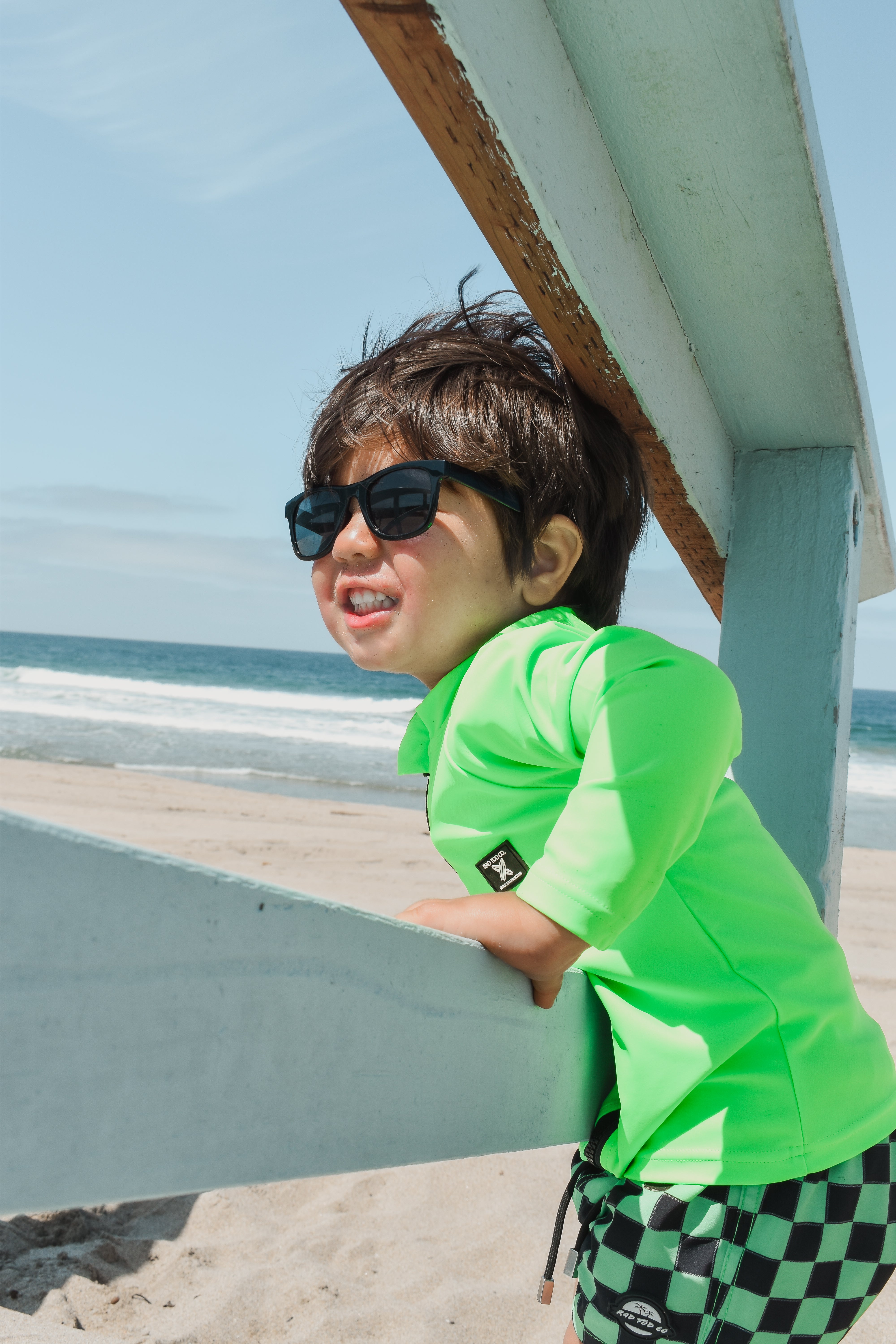 Boy smiling while wearing bright neon zip-front rashguard, protected from sun while easily visible against sandy beach background.