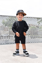 Child wearing a black outfit with a cap, standing on a concrete surface with a metal fence background.