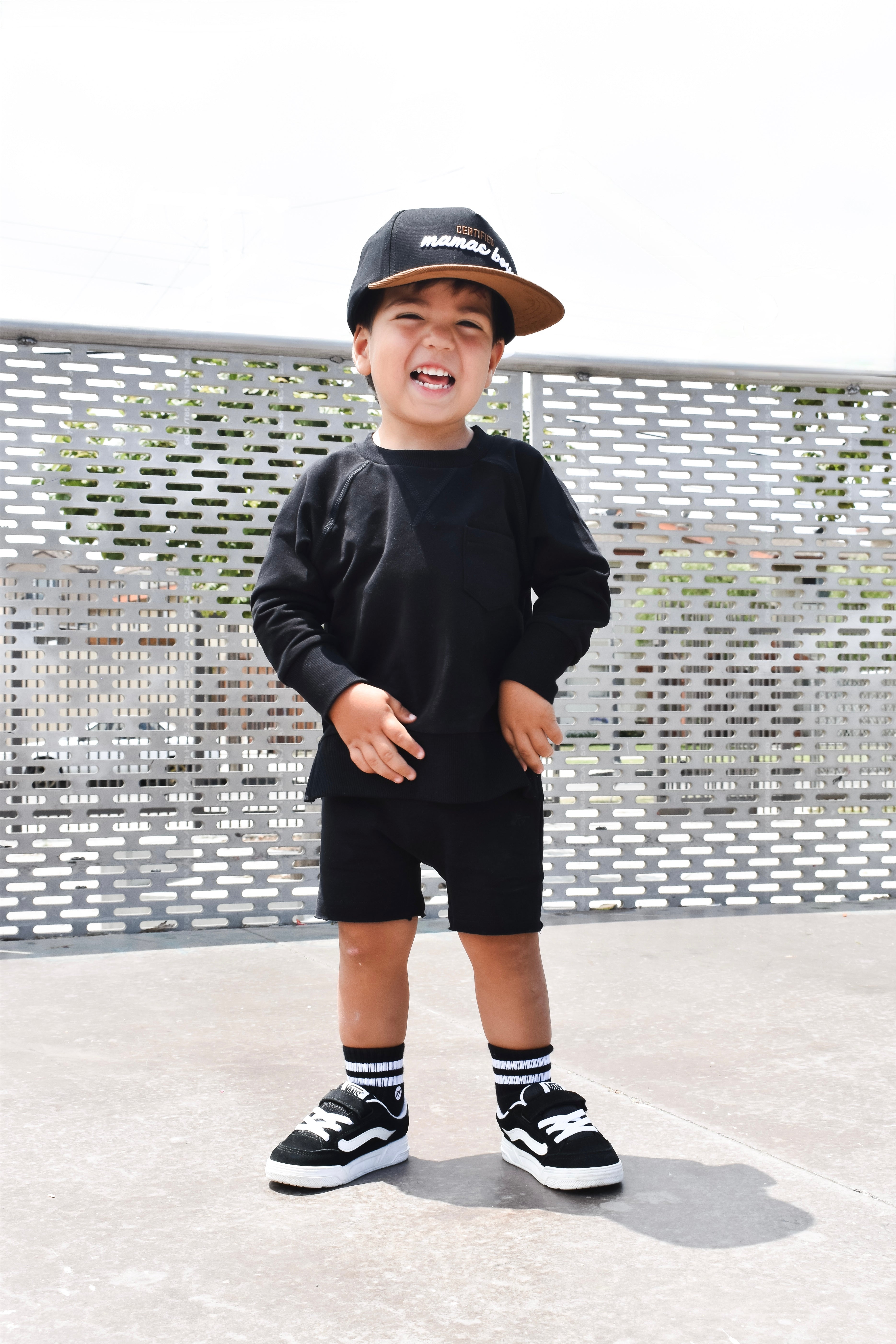 Child wearing a black outfit with a cap, standing on a concrete surface with a metal fence background.
