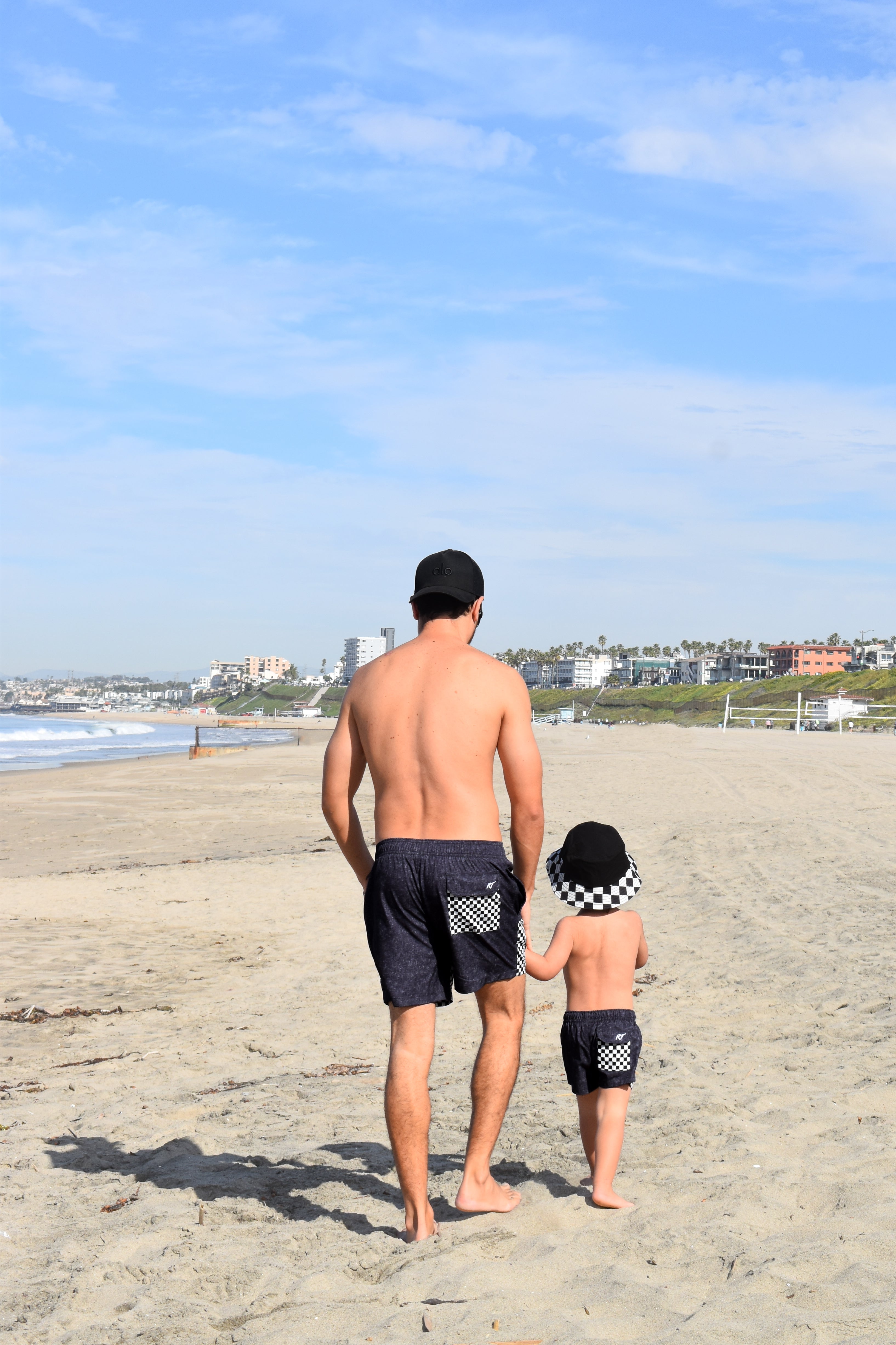 Man and child walking on a beach with a clear blue sky wearing matching board shorts