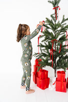 Child in festive pajamas decorating a Christmas tree with red ribbons and presents while wearing ornament lane bamboo by RAD TODDLER