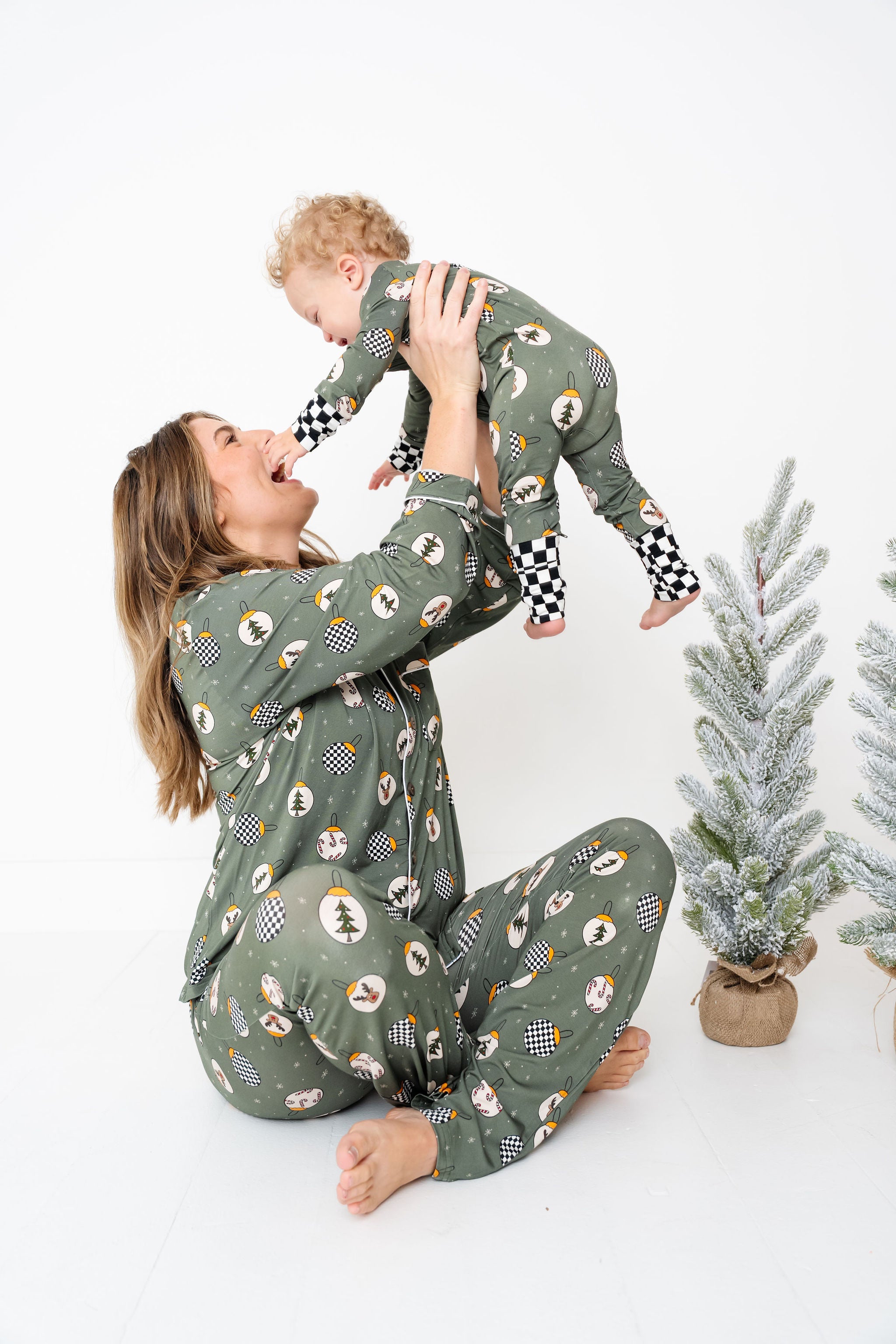 Woman and child wearing matching green pajamas with a pattern by RAD TODDLER, sitting on a white floor with small Christmas trees.
