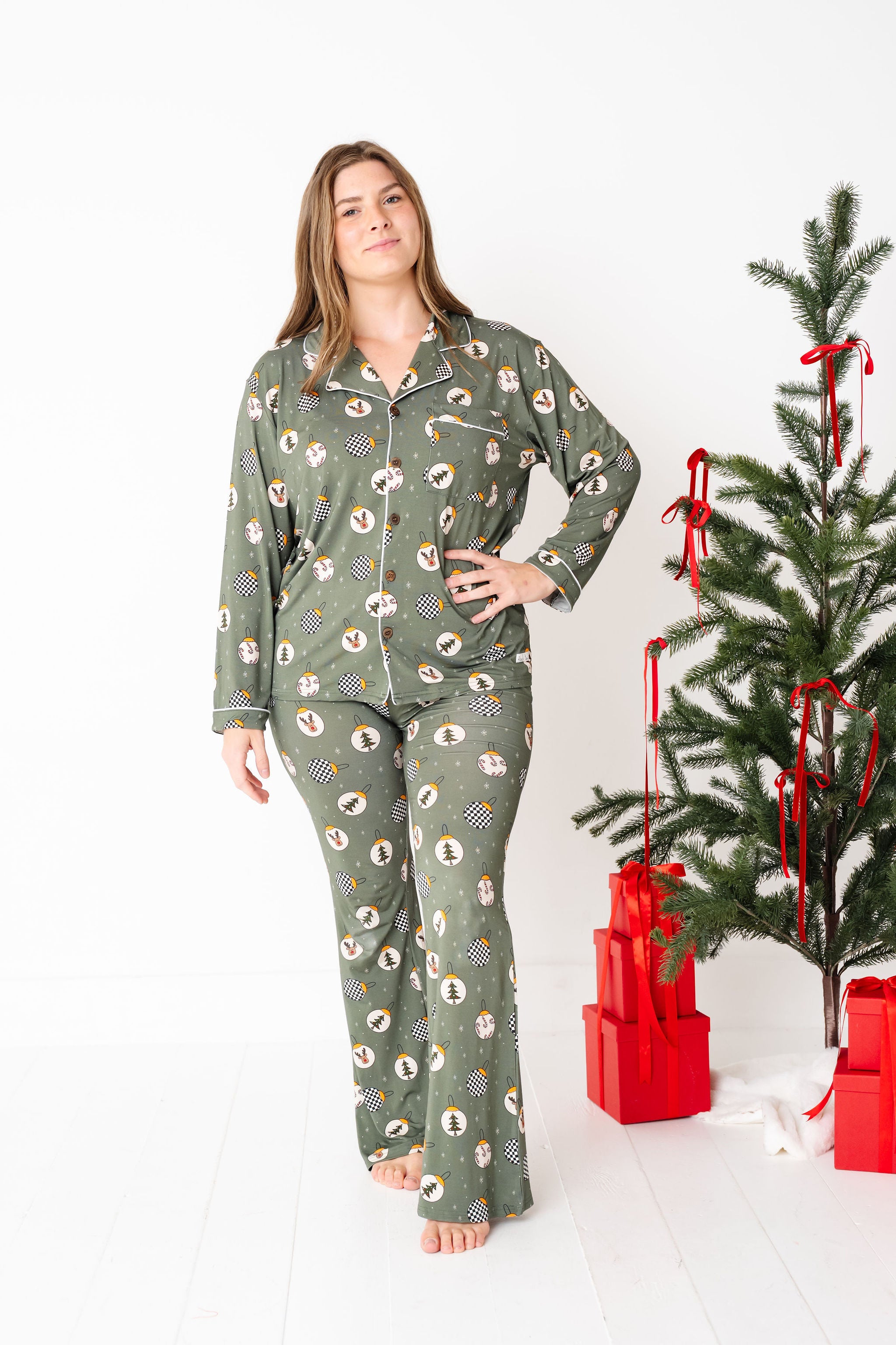 Woman wearing green patterned pajamas standing next to a decorated Christmas tree with red bows and presents.