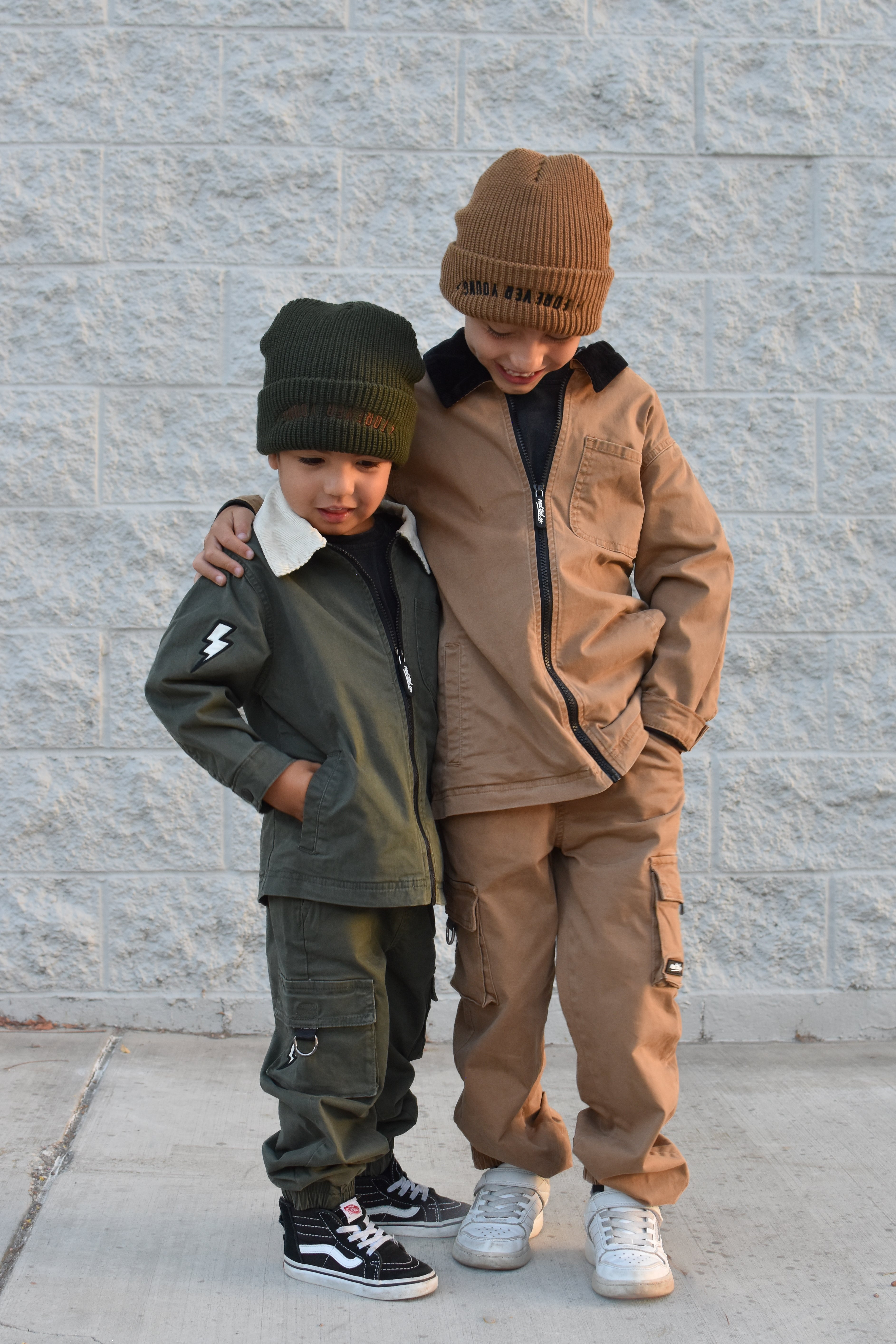 Two children standing outdoors against a light gray brick wall, wearing matching streetwear outfits. The child on the left wears an olive green jacket and pants with a dark green beanie, and the child on the right wears a tan/brown jacket and matching pants with a brown beanie.