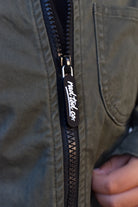 Close-up of a black zipper on an olive green jacket, featuring a black pull tab with white script text reading 'RAD TOD CO.' and a hand holding the zipper open.