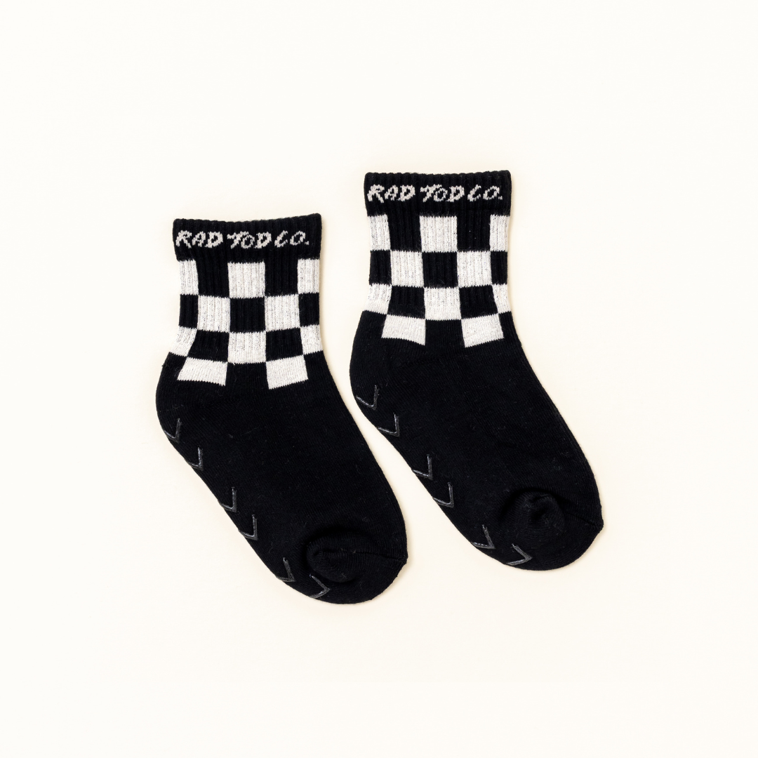 A pair of black crew socks with a white checkerboard pattern around the cuffs and the brand name 'ROD TOD CO.' printed in white on the top of each cuff, displayed on a clean off-white background.