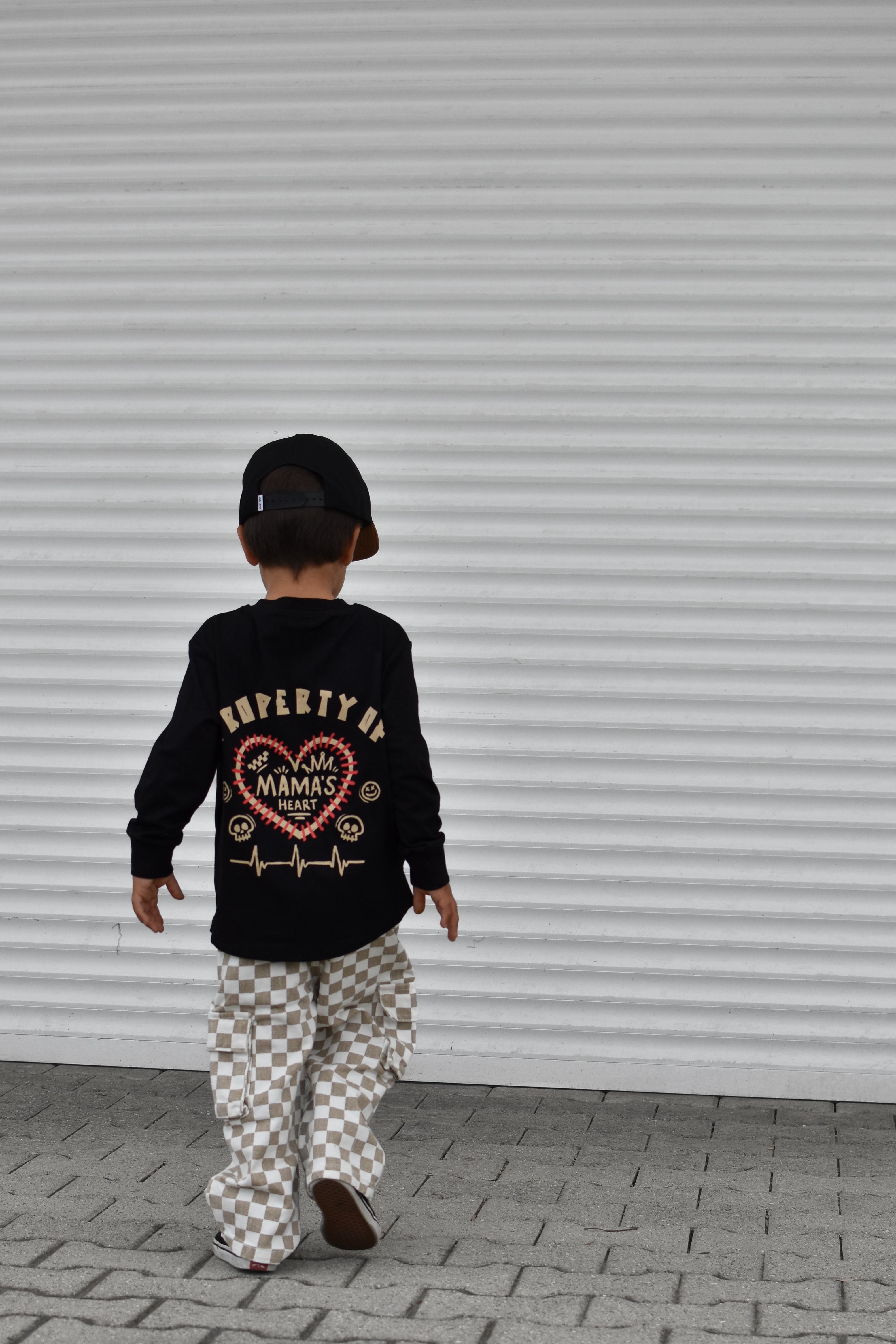 A child seen from behind wearing a black long-sleeve graphic sweatshirt with a heart design that reads 'PROPERTY OF MAMAS HEART' and checkered pants, walking in front of a white corrugated metal shutter.