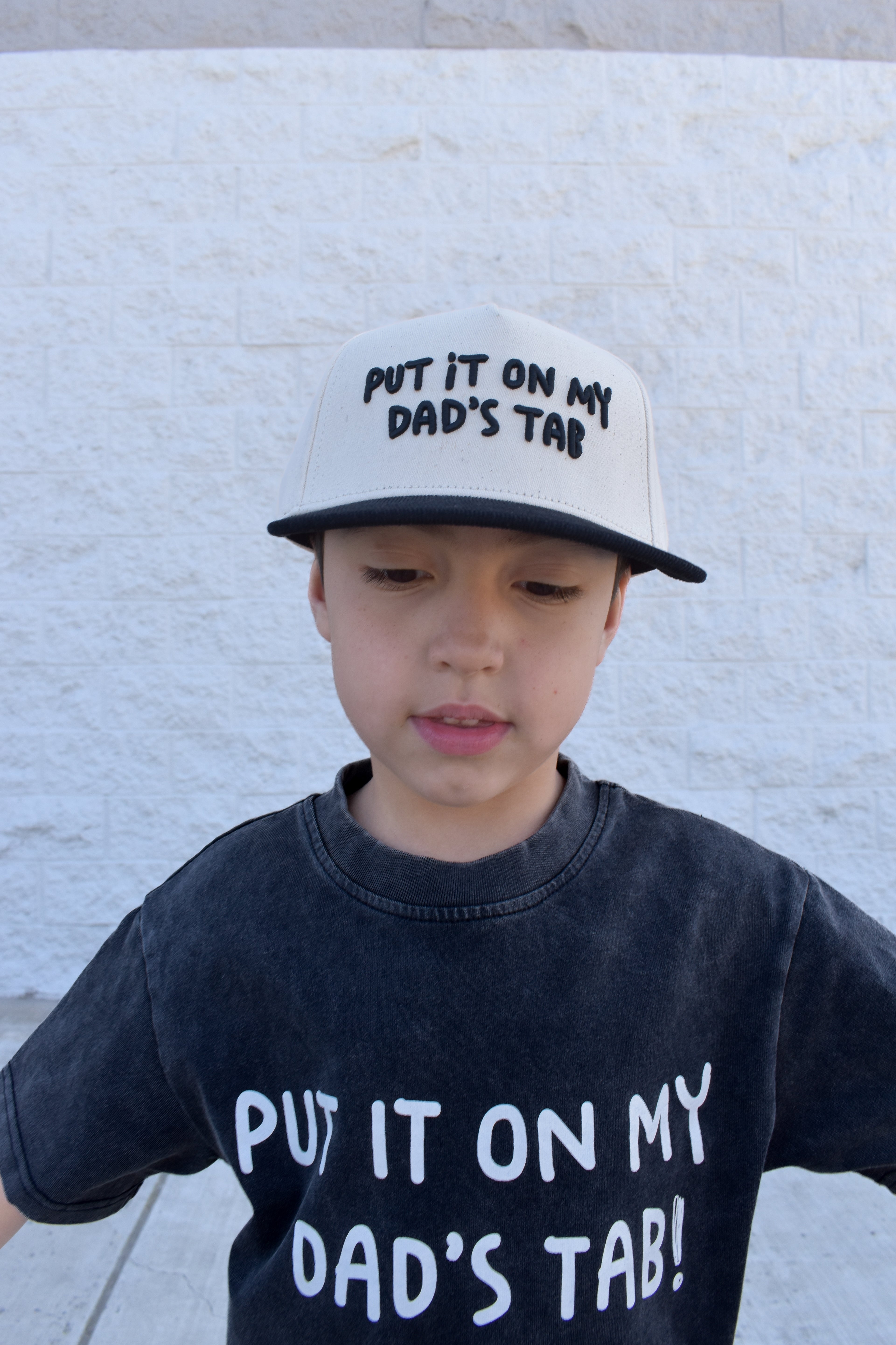 Child wearing a cap and t-shirt with Put It On My Dad’s Tab against a light-colored wall.