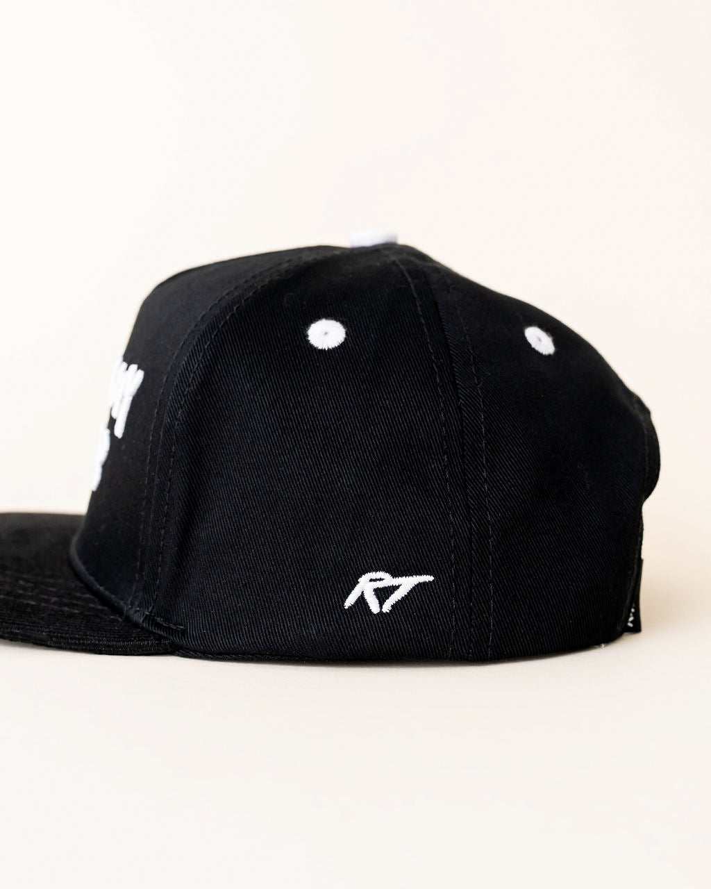 A black snapback cap shown from a side angle, featuring a white embroidered stylized logo on the front side panel and a matching white logo on the brim, set against a plain off-white background.