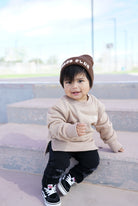 Child wearing a beige sweatshirt, black pants, and a brown beanie sitting on steps.
