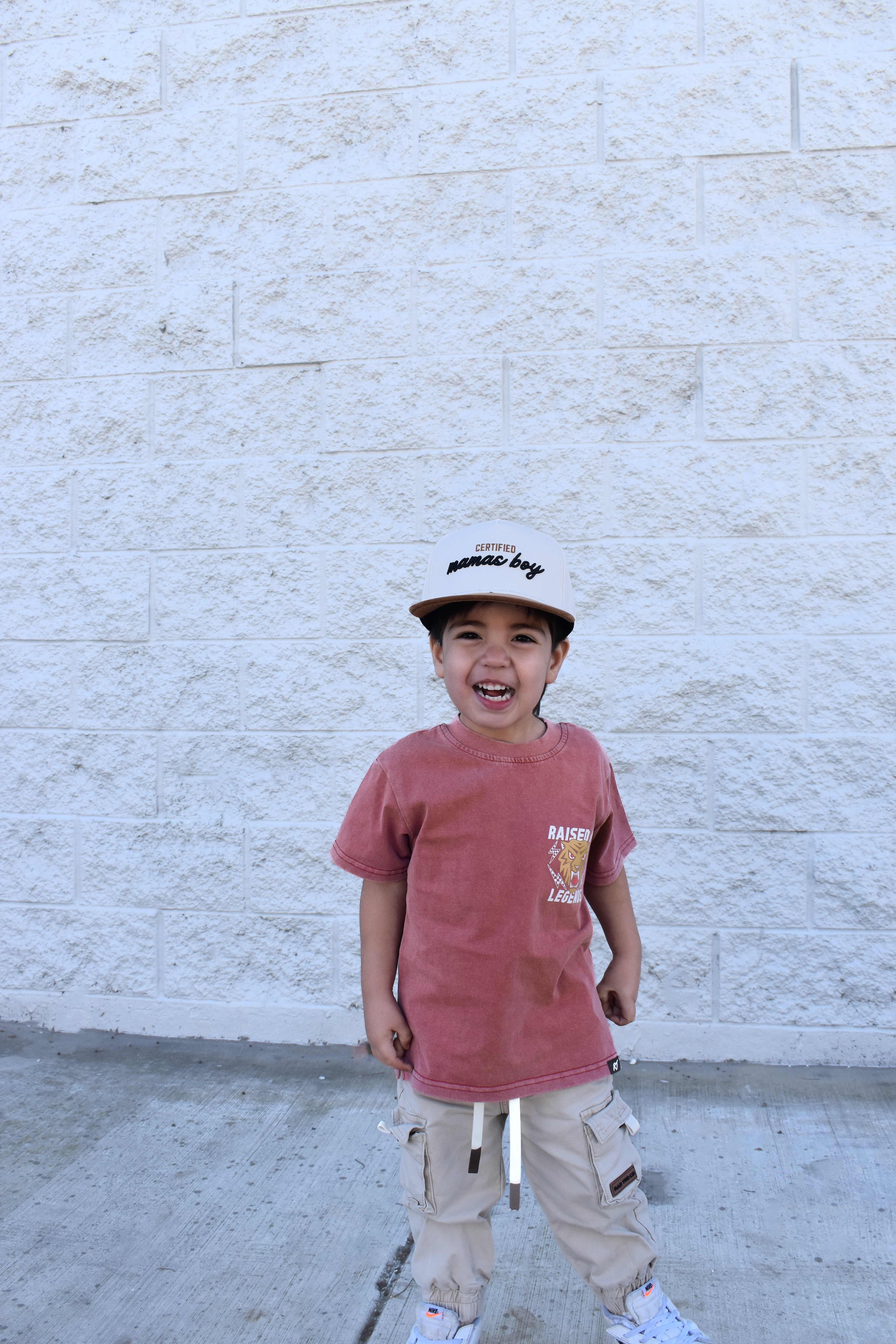 Child wearing a red shirt and beige cap standing against a white brick wall.