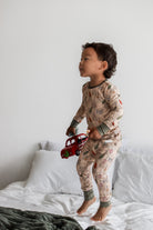 A toddler stands barefoot on a bed wearing a beige patterned long-sleeve pajama set with green cuffs and holding a red toy truck.
