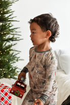 A toddler wearing a holiday-themed long-sleeve pajama set with a Christmas tree and ornament print, standing next to a decorated Christmas tree and holding a small red toy truck.