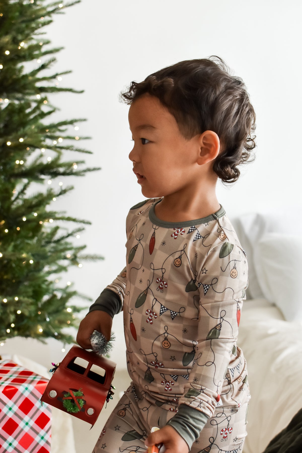 A toddler wearing a holiday-themed long-sleeve pajama set with a Christmas tree and ornament print, standing next to a decorated Christmas tree and holding a small red toy truck.