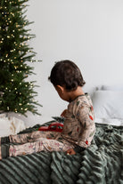 A toddler wearing a beige Christmas-themed long-sleeve pajama set with a holiday lights print, sitting on a dark green blanket next to a decorated Christmas tree.