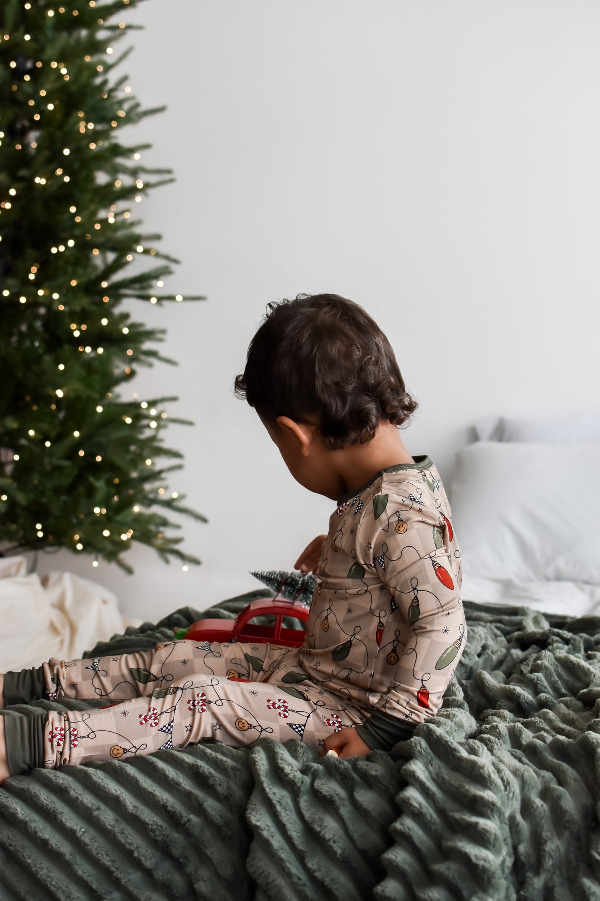 A toddler wearing a beige Christmas-themed long-sleeve pajama set with a holiday lights print, sitting on a dark green blanket next to a decorated Christmas tree.
