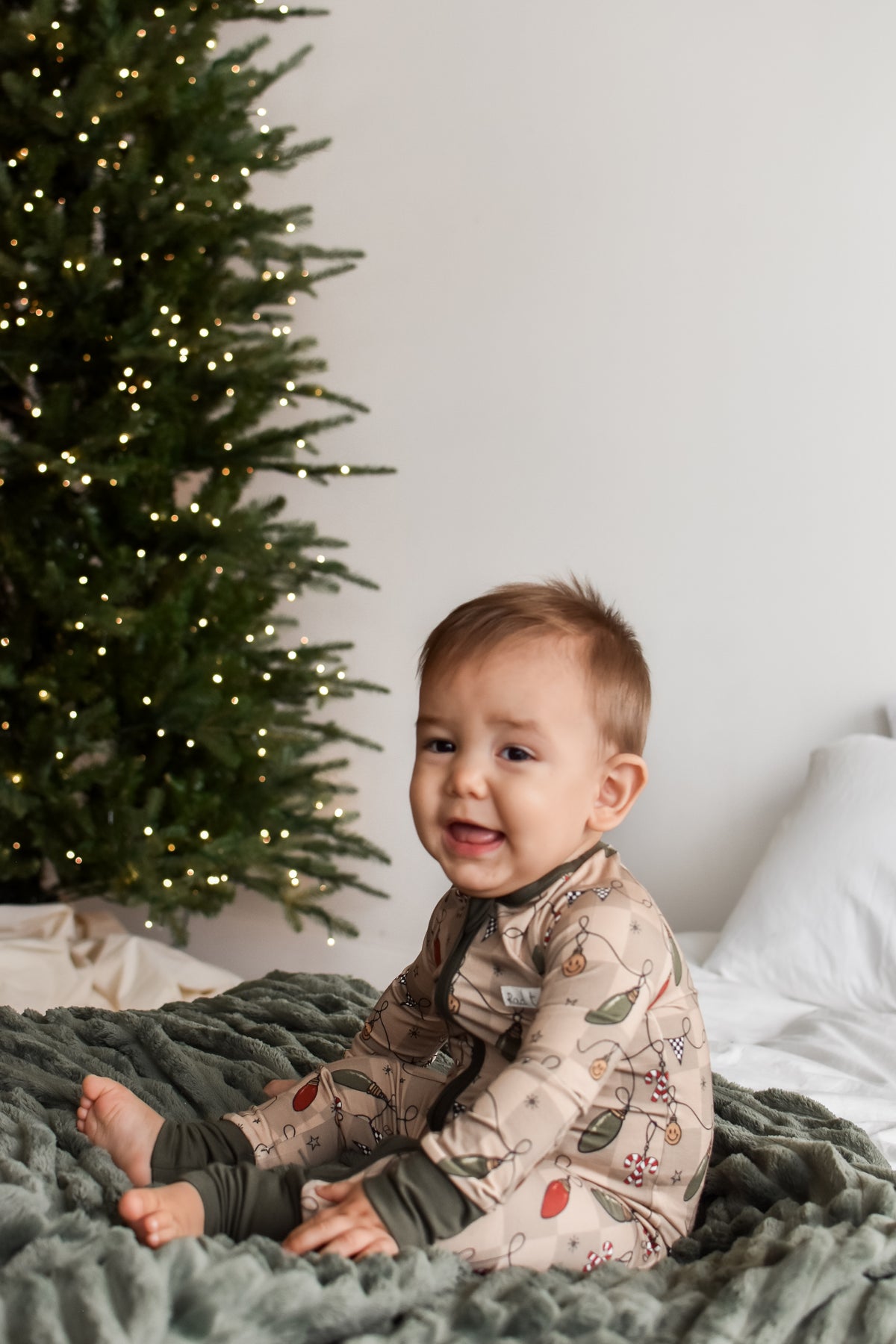 A baby sitting on a green blanket in front of a Christmas tree, wearing a beige long-sleeve pajama with a winter botanical pattern.