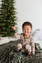 A smiling young child wearing a festive holiday pajama set  sitting on a ribbed dark green blanket next to a decorated Christmas tree.