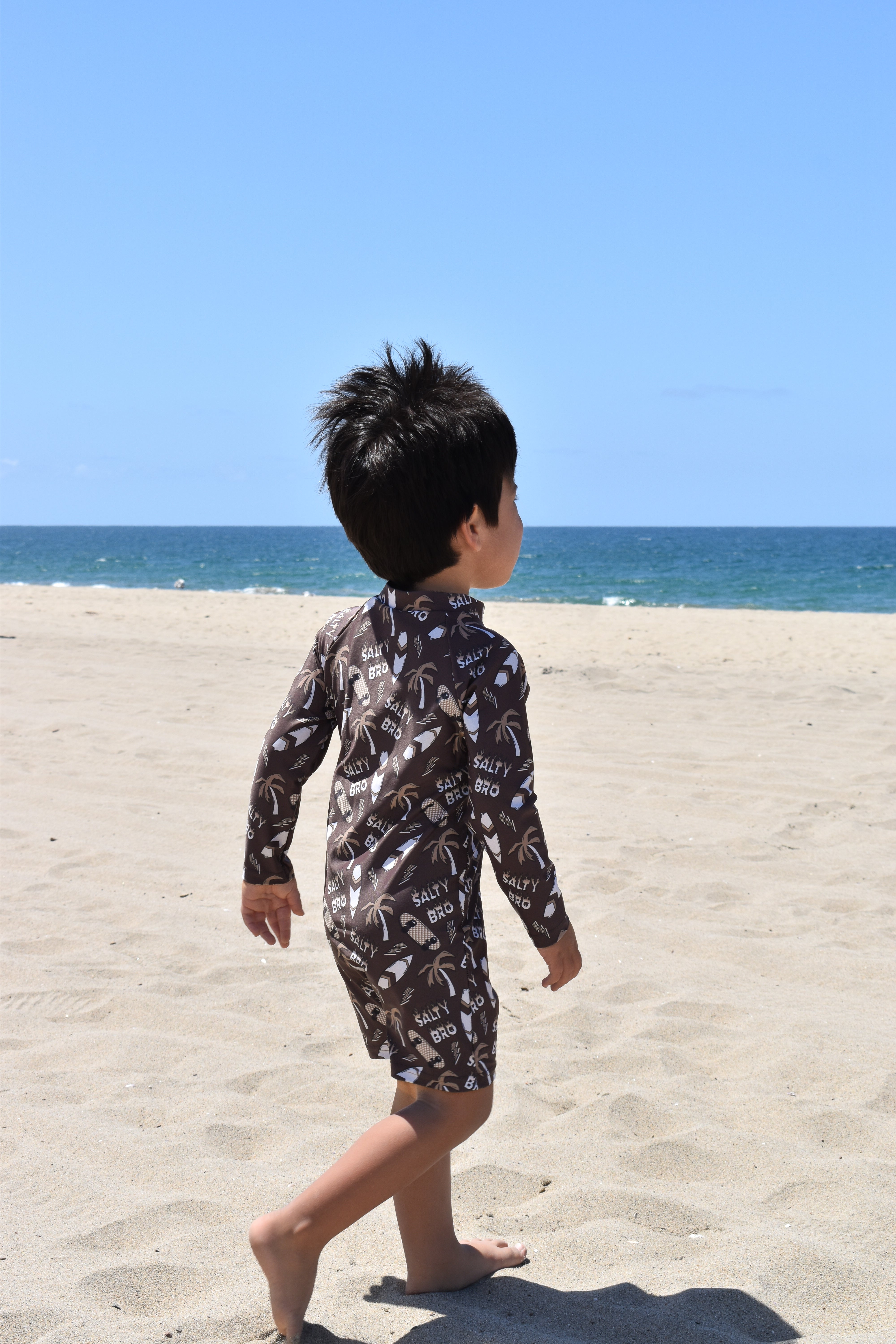 Child in a patterned swimsuit walking on a sandy beach with clear blue sky and ocean.