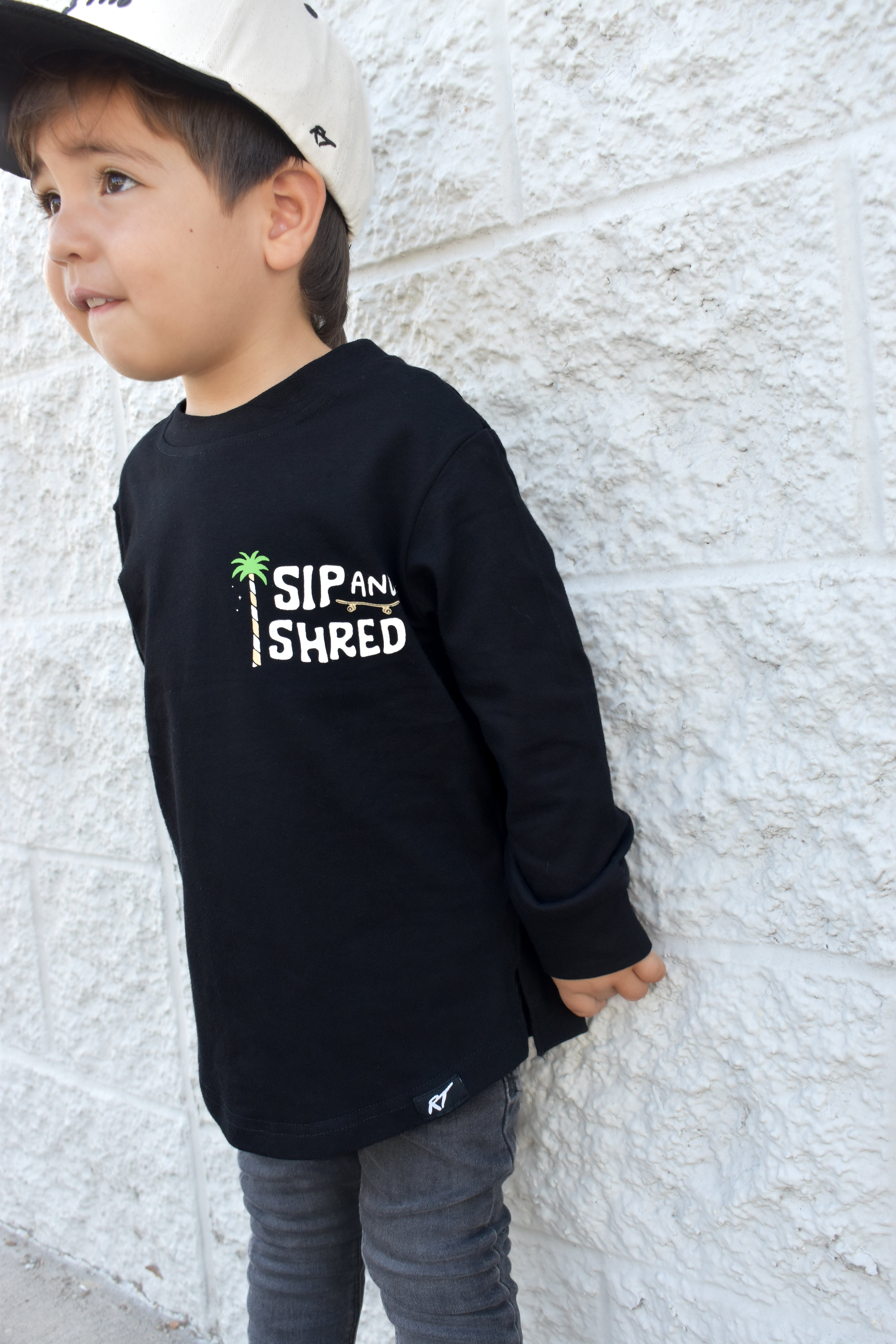 Child wearing a black sweatshirt with 'Sip and Shred' text against a light stone wall.