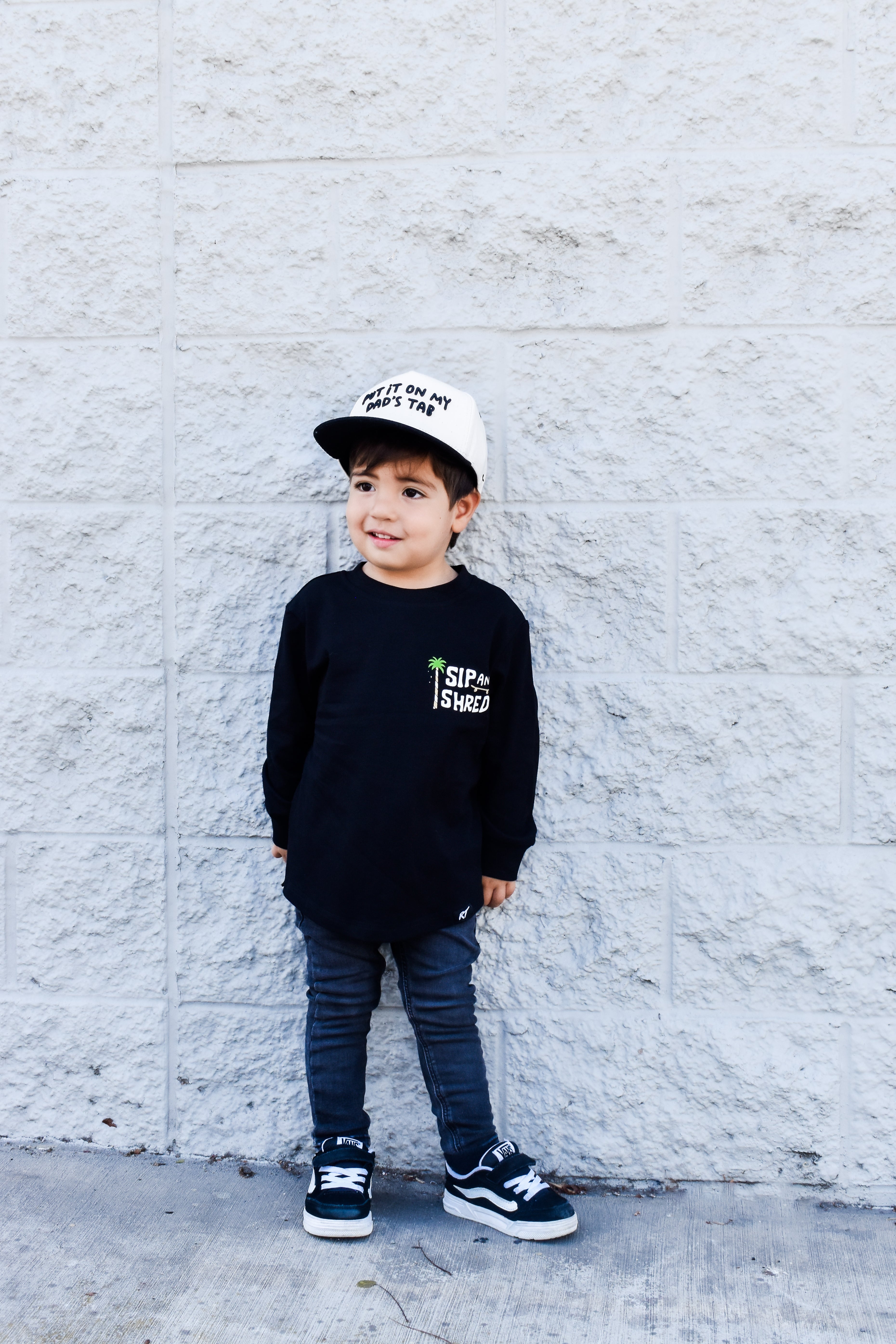 Child wearing a black sweatshirt with text, blue jeans, and a white cap against a light gray brick wall.