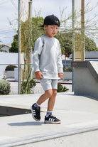 Child in gray sweatshirt and shorts walking on a concrete surface with trees and buildings in the background