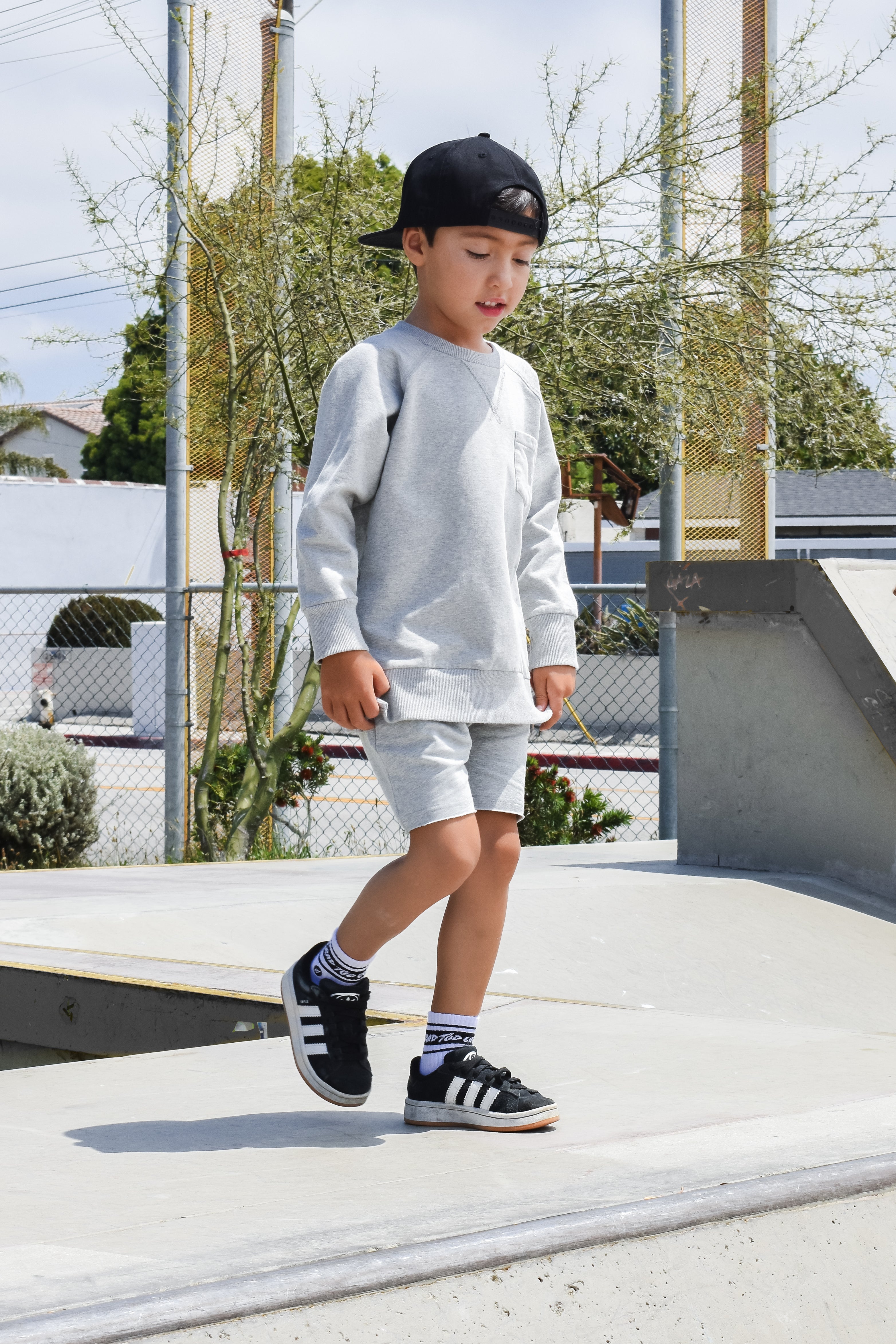 Child in gray sweatshirt and shorts walking on a concrete surface with trees and buildings in the background