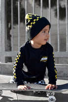 A child wearing a black and yellow checkered beanie and a black long-sleeve shirt with yellow accents sits on a skateboard in front of a gray metal fence.