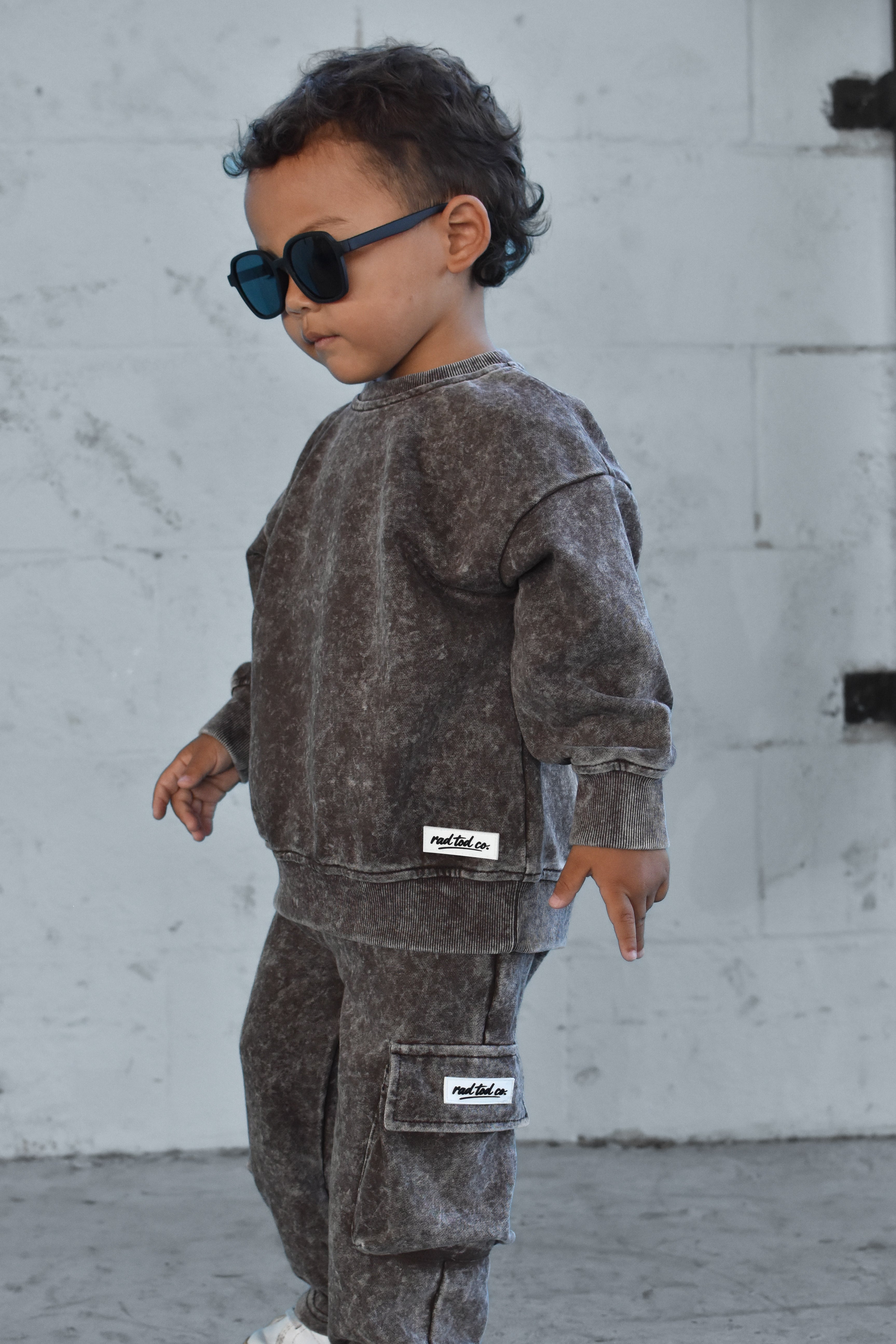 A toddler wearing a matching washed dark brown sweatshirt and cargo pants set with sunglasses, standing against a light gray concrete wall.
