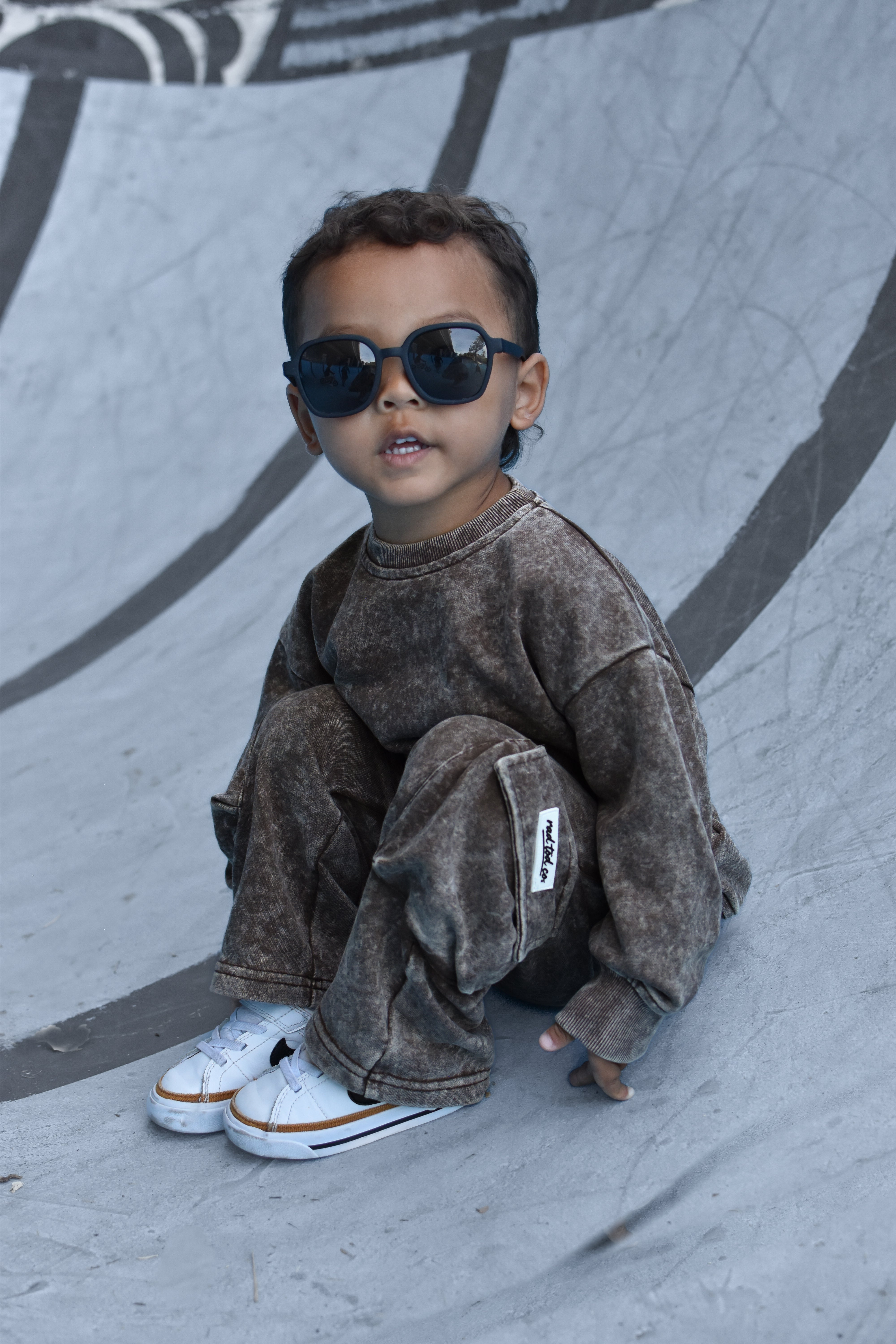 A toddler wearing a matching brown acid-wash sweatshirt and jogger pants set, styled with black sunglasses and white sneakers, sitting on a concrete skatepark ramp.
