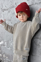 A toddler wearing a beige crewneck sweatshirt and matching pants with a red knit beanie, posing against a textured gray concrete wall.