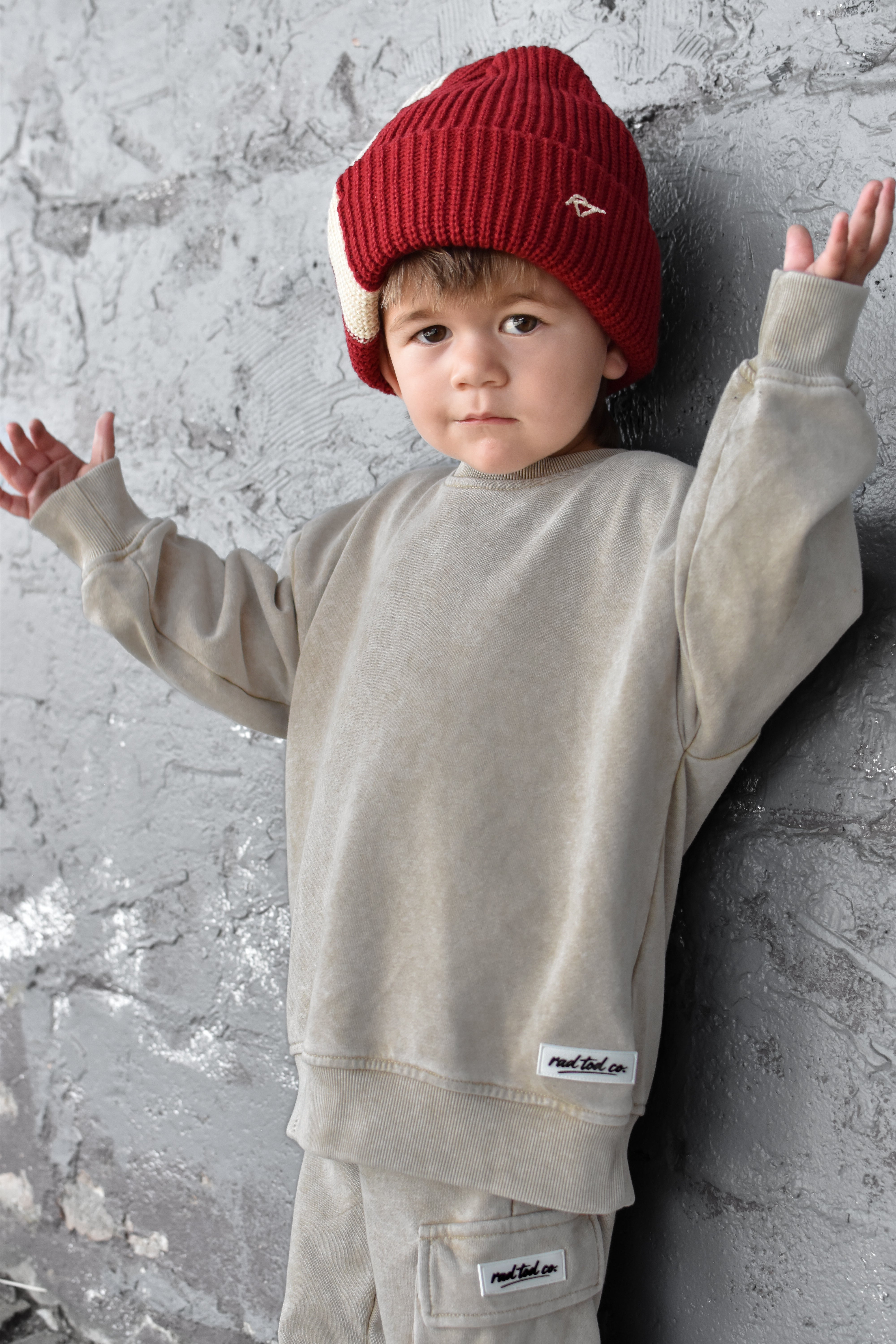 A toddler wearing a beige crewneck sweatshirt and matching pants with a red knit beanie, posing against a textured gray concrete wall.