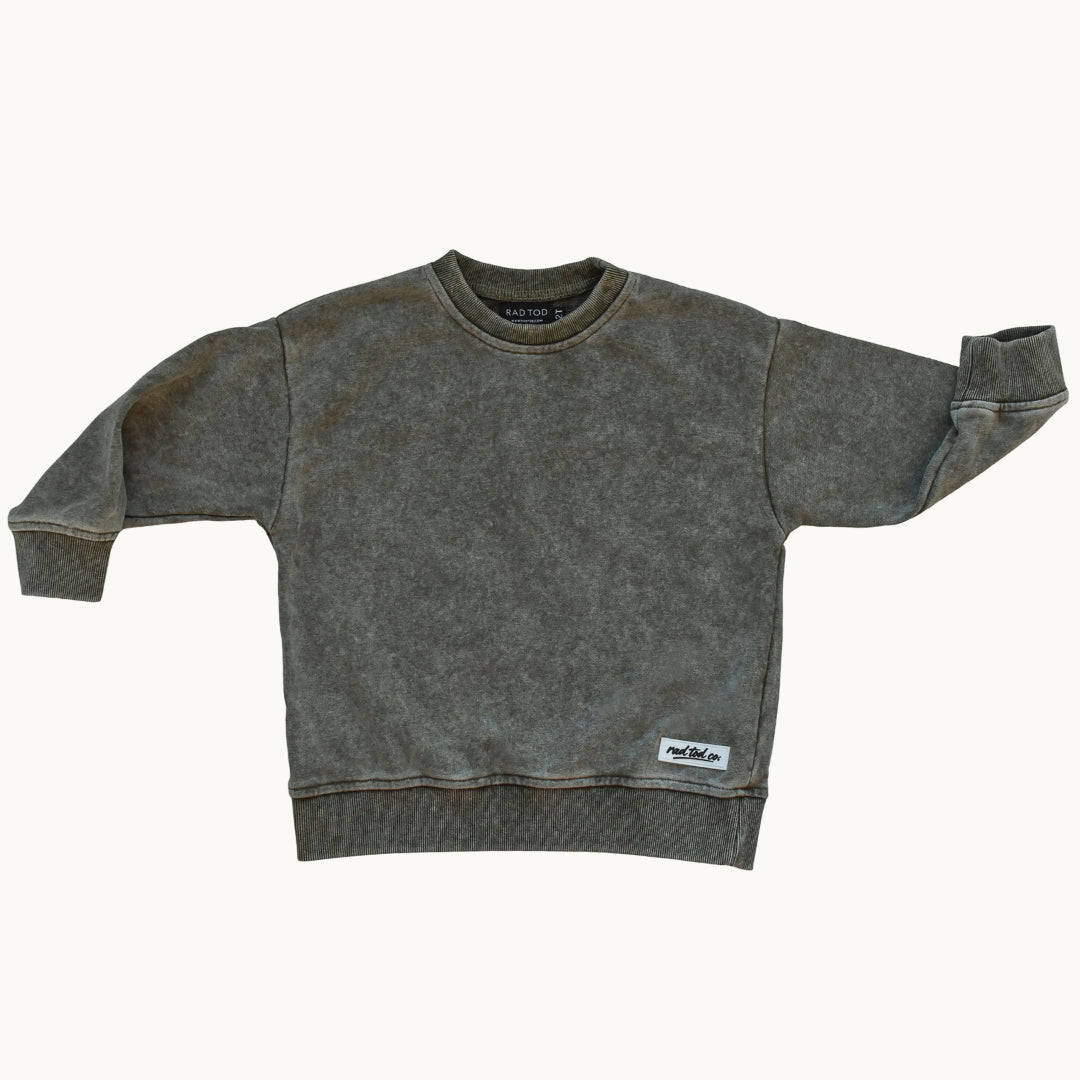 A stone washed moss sweatshirt for kids, laid flat against a light off-white background. The sweatshirt features a crew neck, ribbed cuffs, and a small white brand patch on the lower right hem.
