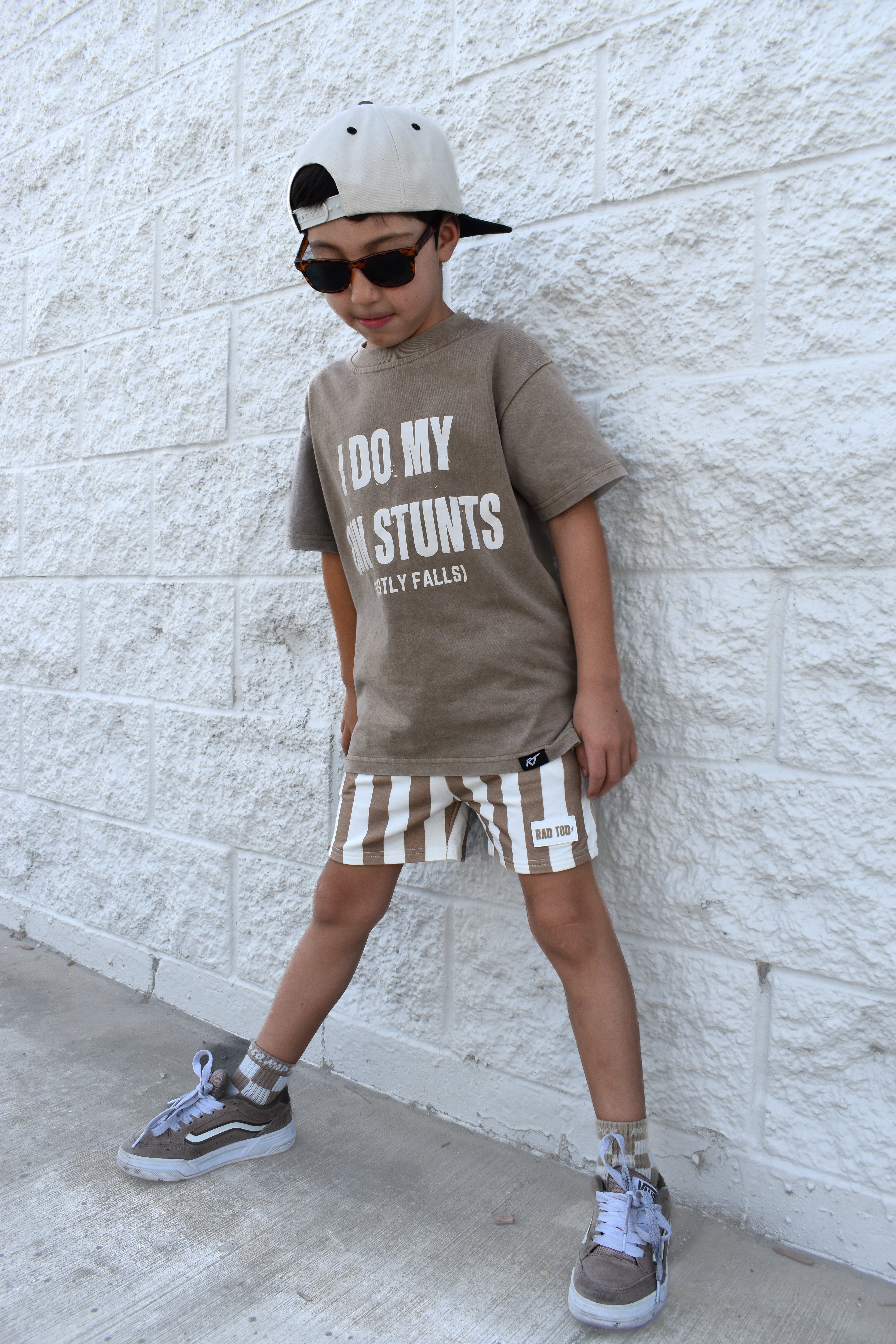 Child wearing a brown t-shirt with text, striped shorts, and sunglasses against a white brick wall.