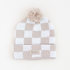 A checkered knit beanie hat with a pom-pom on top and a folded cuff, featuring a small brand label that reads 'RAD TOD CO' and 'WINTERTIDE COLLECTION'. The colors are a warm taupe-beige and white, set against a clean off-white background.