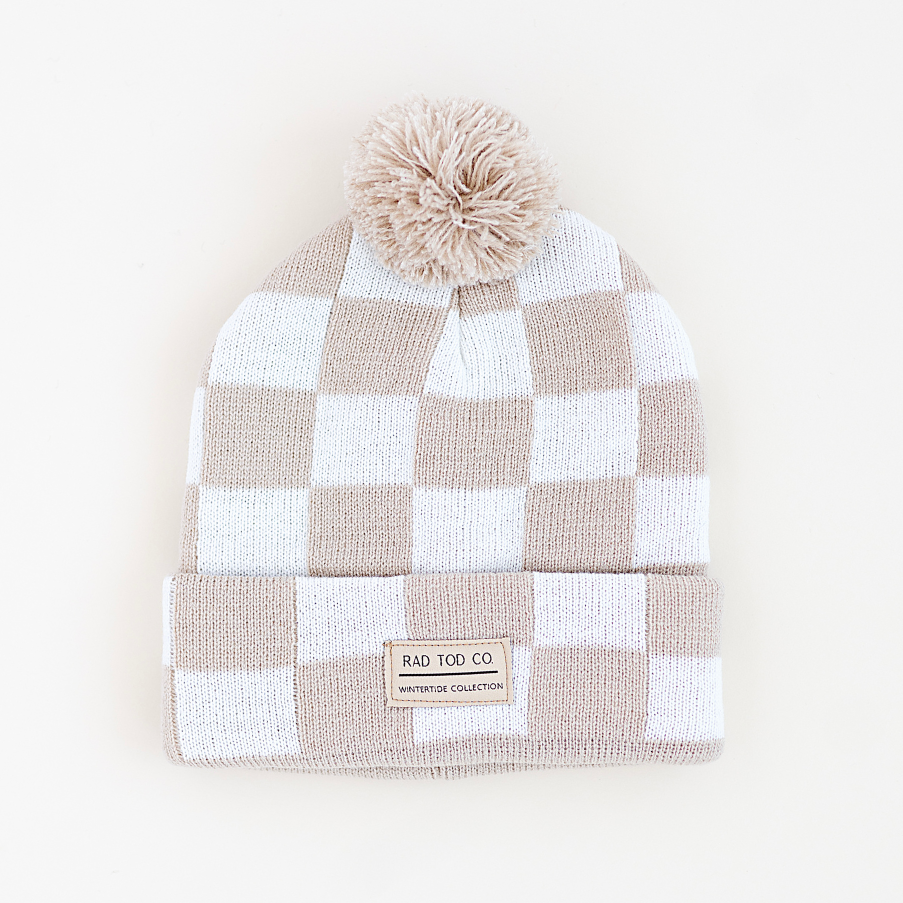 A checkered knit beanie hat with a pom-pom on top and a folded cuff, featuring a small brand label that reads 'RAD TOD CO' and 'WINTERTIDE COLLECTION'. The colors are a warm taupe-beige and white, set against a clean off-white background.