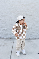 Child wearing a checkered jacket and cargo pants standing against a light gray brick wall.