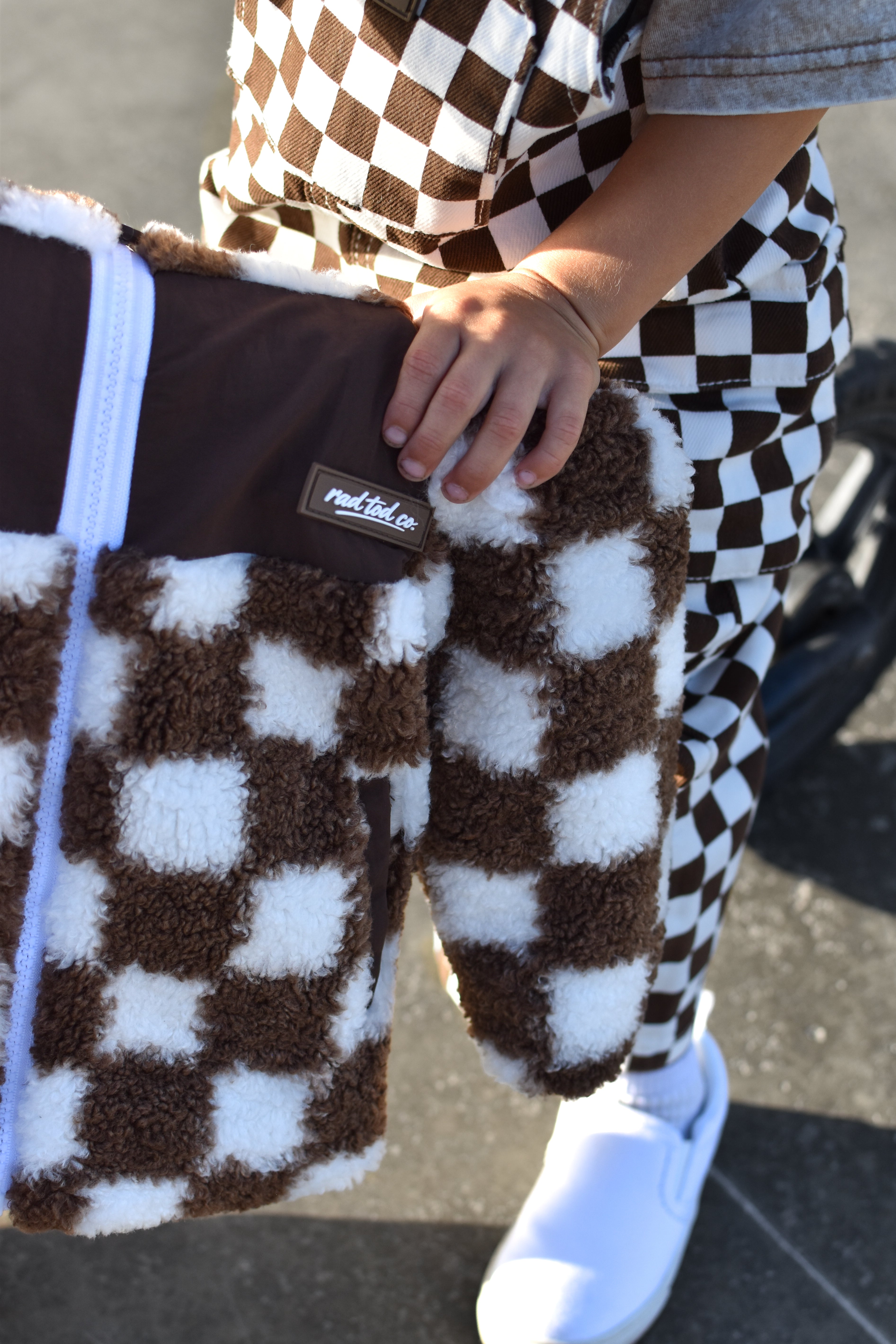 A close-up of a person holding a brown and white checkered teddy sherpa jacket outdoors. The person is wearing matching brown and white checkered pants, white slip-on shoes, and a gray shirt