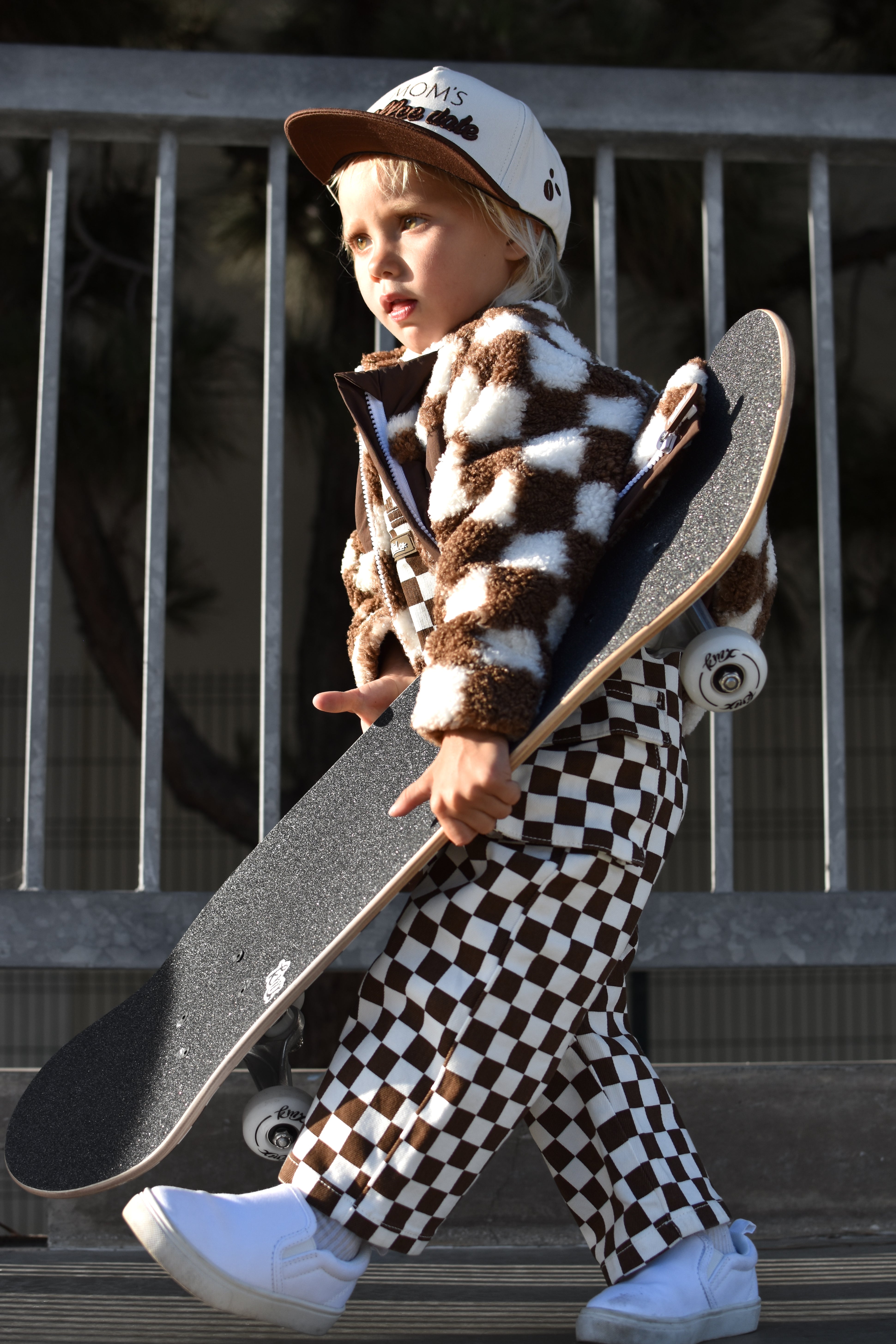 A toddler walking while carrying a skateboard, wearing a brown and white checkered fuzzy jacket and matching checkered pants, a white cap, and white sneakers.
