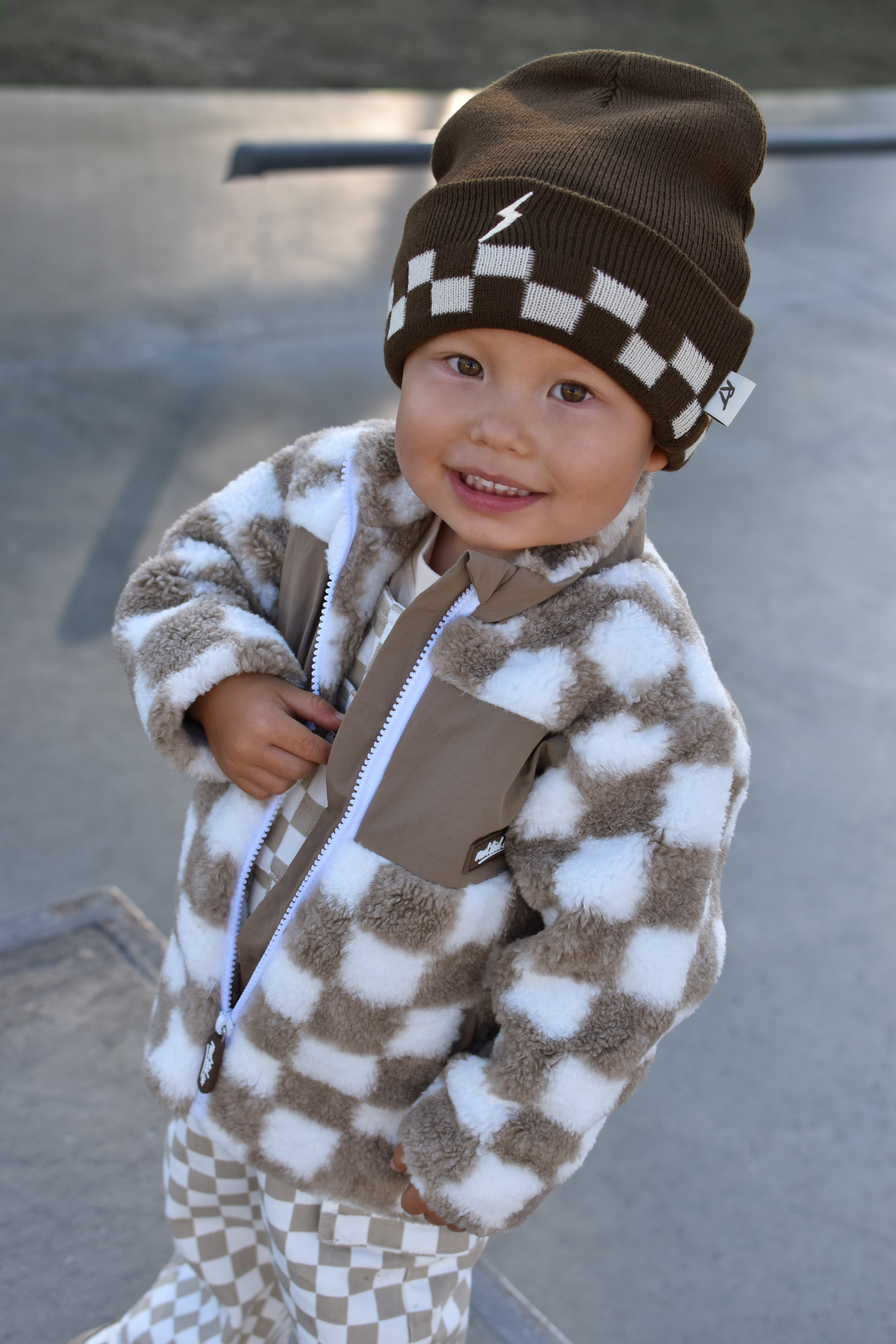 A smiling toddler wearing a brown and white checkered fuzzy zip-up jacket, matching checkered pants, and a brown beanie with a white lightning bolt, standing outdoors on a concrete surface.