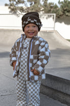 A smiling toddler standing outdoors in a skatepark, wearing a brown and beige checkered fuzzy jacket, matching checkered pants, and a brown beanie with a checkerboard pattern.