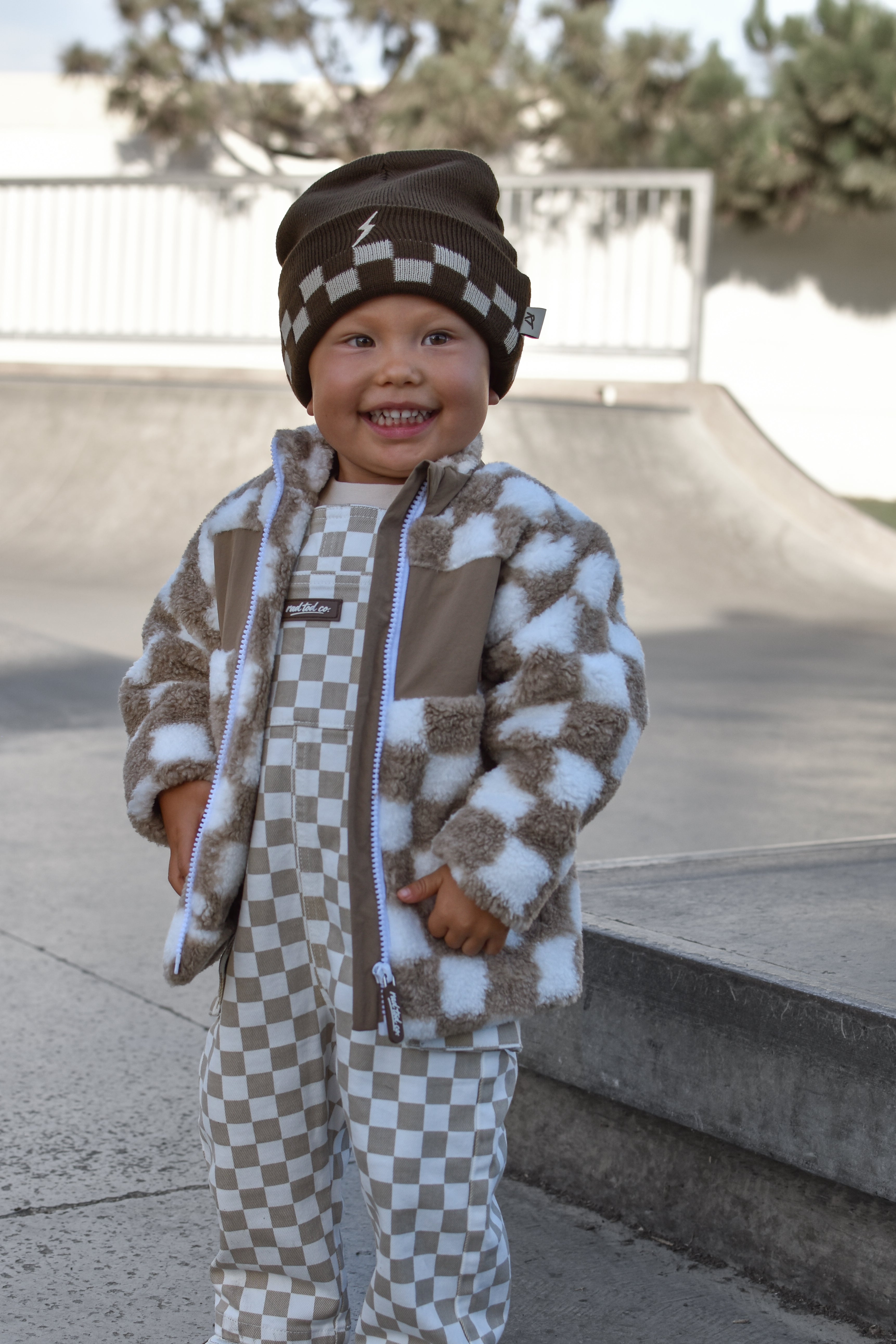 A smiling toddler standing outdoors in a skatepark, wearing a brown and beige checkered fuzzy jacket, matching checkered pants, and a brown beanie with a checkerboard pattern.