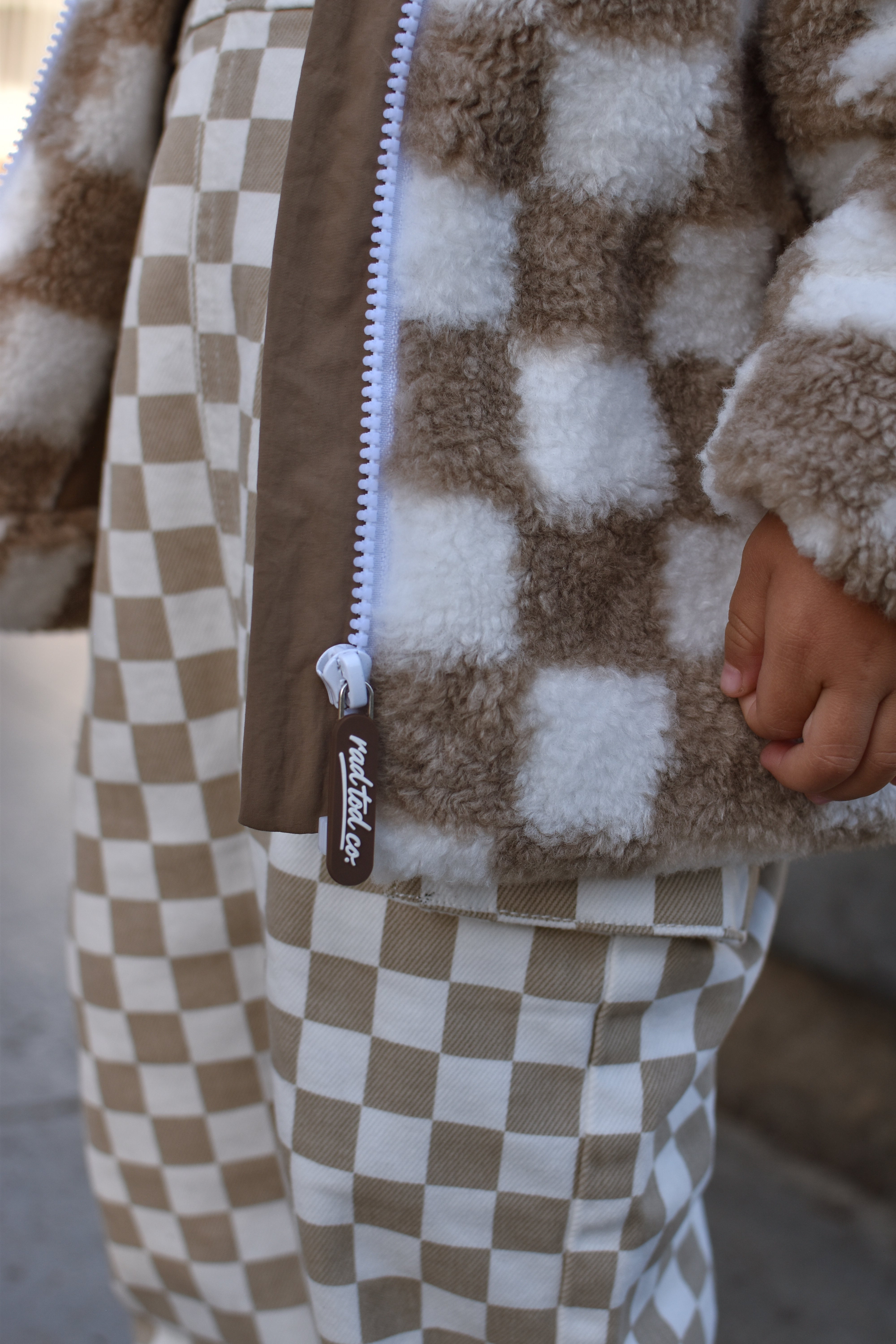 Close-up of a child wearing a brown and cream checkered fleece zip-up jacket and matching checkered pants, with a hand visible at the side.