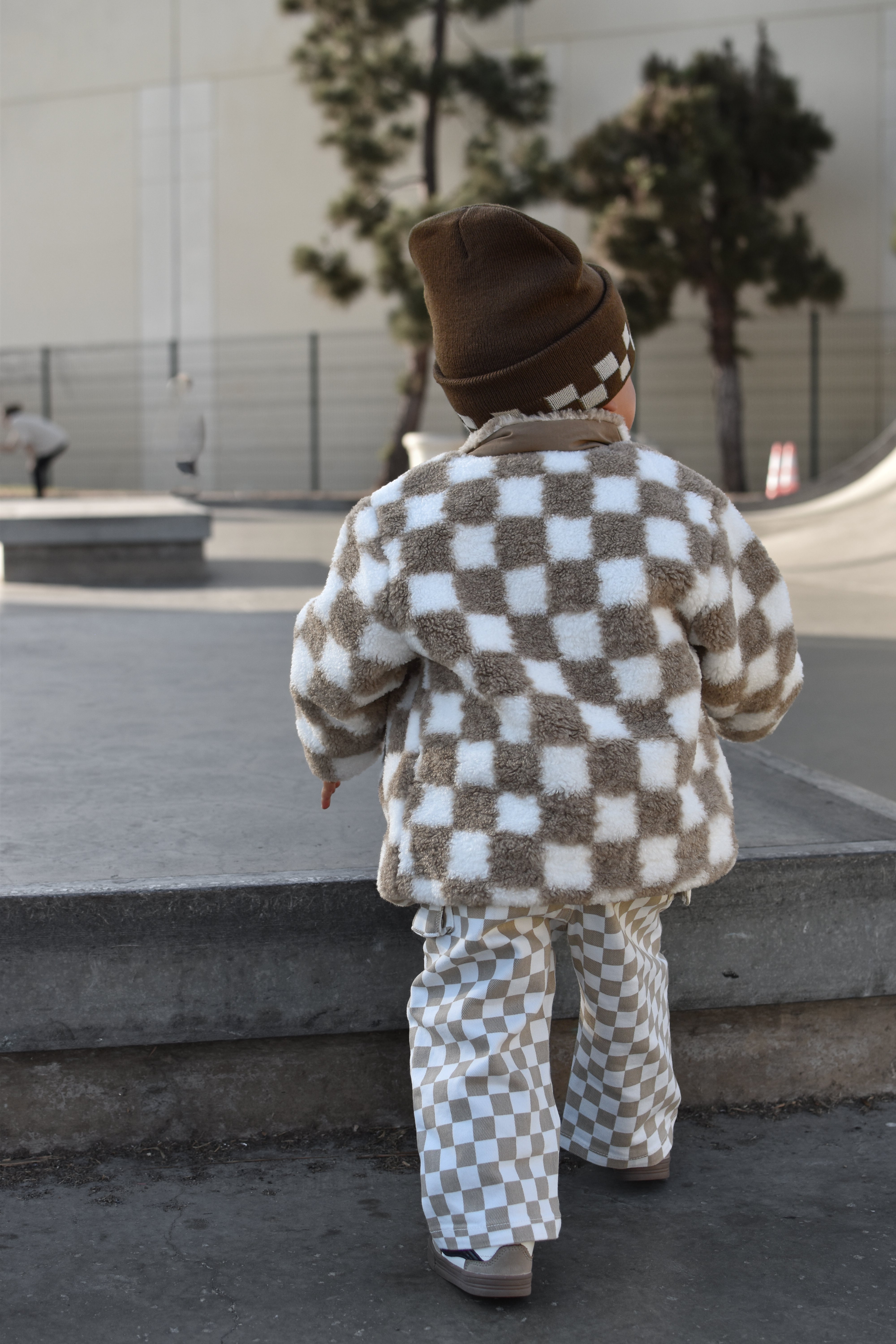 A toddler seen from behind wearing a brown and white checkered fuzzy jacket and matching pants, walking in an outdoor concrete plaza.