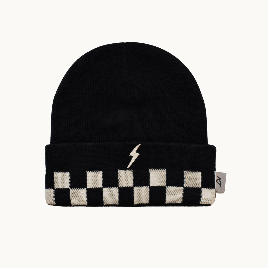 A black knit beanie hat featuring a white lightning bolt emblem on the front of the folded cuff and a black and white checkerboard pattern along the lower edge. A small gray brand tag is visible on the right side of the cuff, set against a solid off-white background.