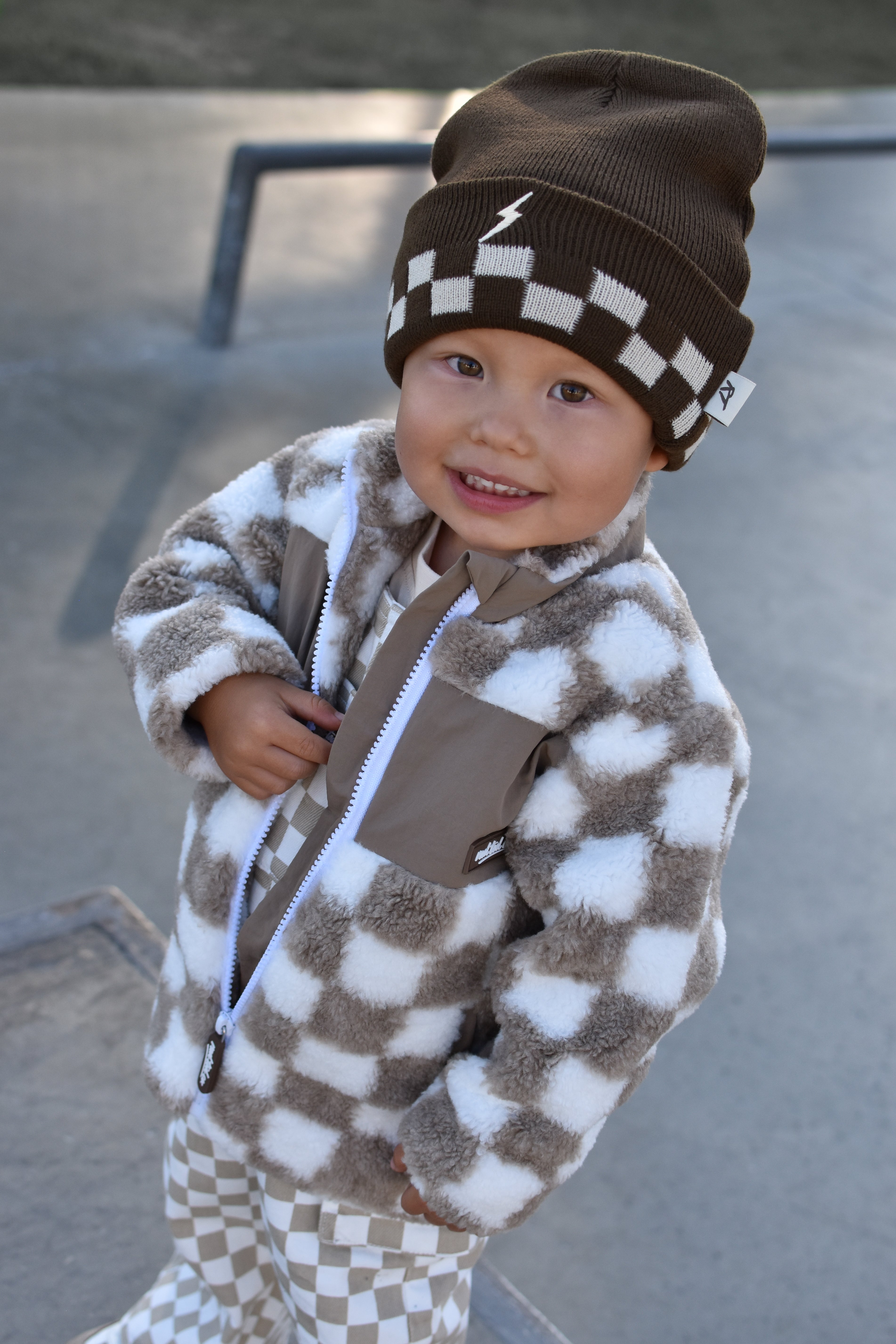 A smiling toddler wearing a brown and white checkered fuzzy zip-up jacket, matching checkered pants, and a brown beanie with a white lightning-bolt patch, photographed outdoors at a skatepark.